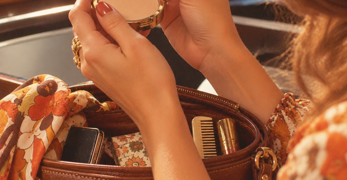 A person holding a compact mirror above an open purse, which contains a comb, lipstick, and floral-patterned items. The person is wearing a floral blouse.