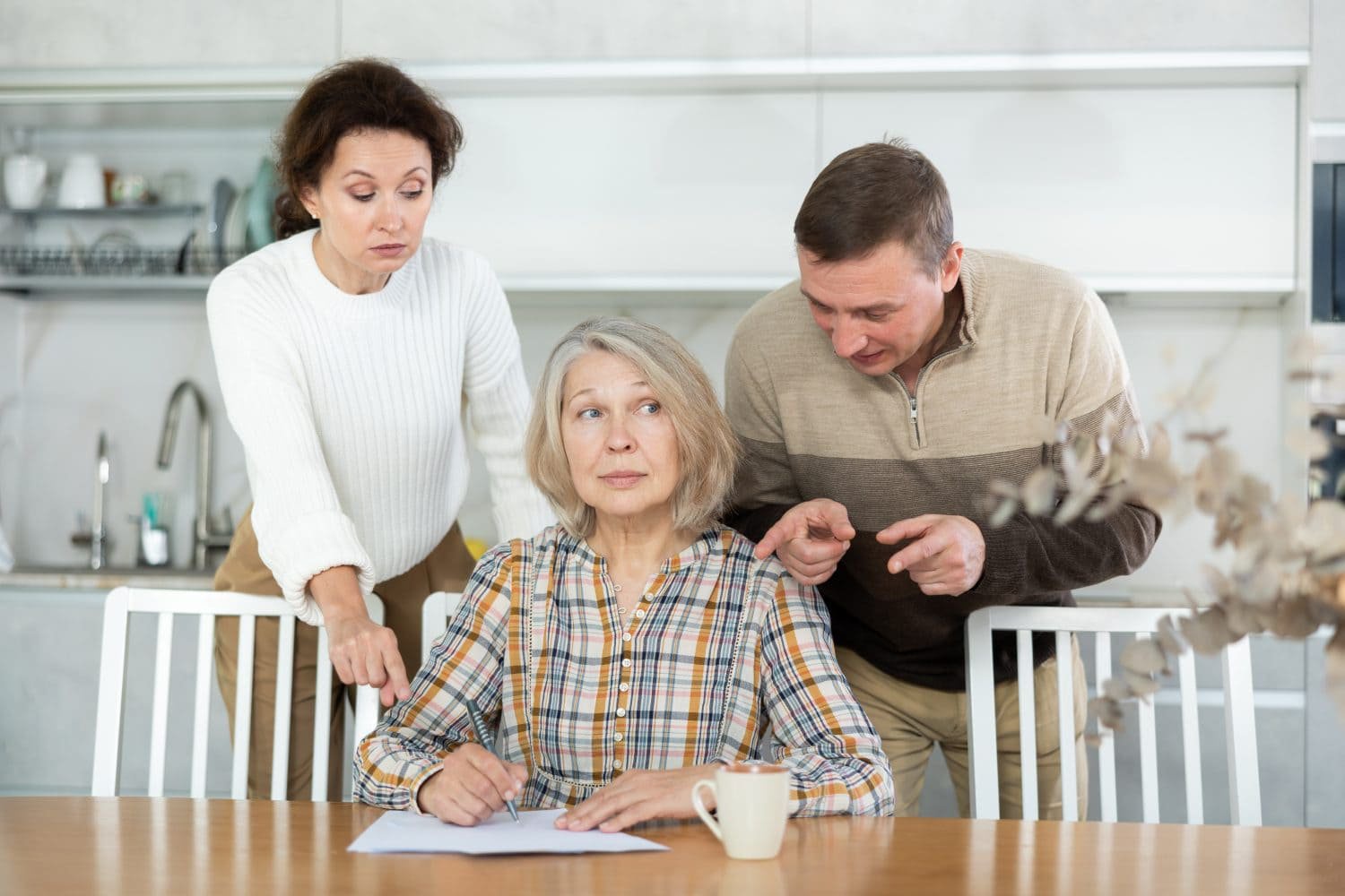 Three individuals are in a kitchen setting. An older woman with gray hair sits at a wooden table, writing on a paper with a pen, looking to her right with a thoughtful expression. A younger woman with dark hair stands behind her to the left, wearing a white sweater, looking down with a concerned face. A man with short brown hair stands behind the older woman to the right, wearing a beige and brown striped sweater, leaning forward and pointing towards the table, seemingly explaining something. A white mug is on the table next to the paper, and a modern kitchen with white cabinets is in the background.