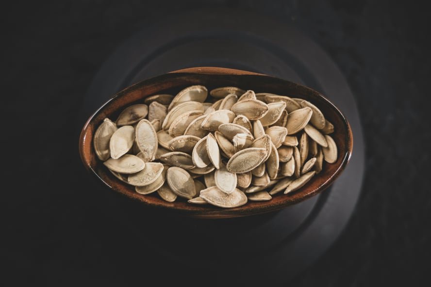 Image of a small brown bowl full of raw pumpkin seeds on a dark counter.