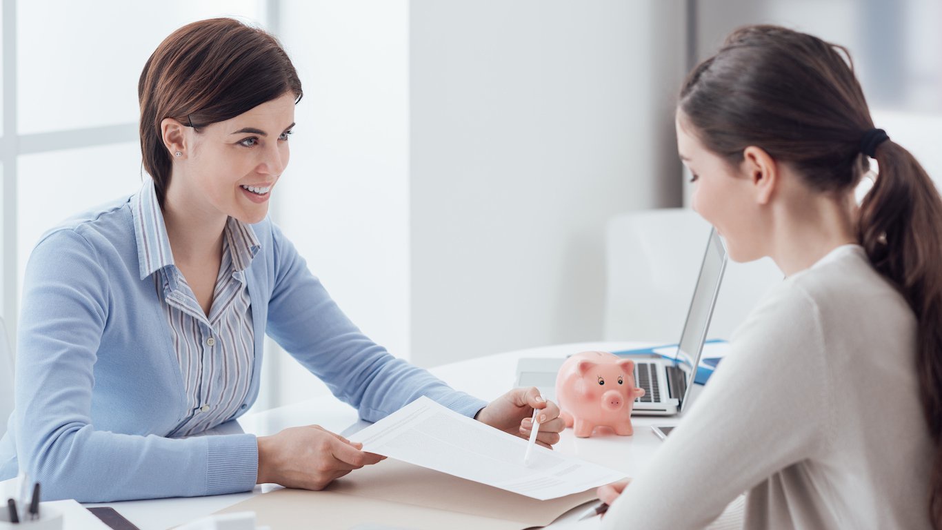 Two women are seated at a white desk in a brightly lit room. The woman on the left, smiling, wears a light blue cardigan over a striped shirt and holds up white papers, gesturing towards the woman on the right. The woman on the right, seen from the side with her hair in a ponytail, looks down at the documents. Between them on the desk, there is a pink piggy bank and a silver laptop.