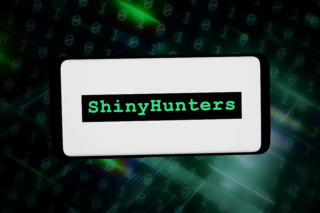 Digital art of a phone with a white screen and a ShinyHunters logo overlaid on it. There are blurry lines of code in the background.