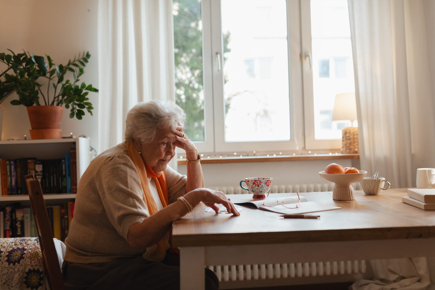 A somber elderly woman with white hair, wearing a beige top and an orange scarf, sits at a wooden table. Her right hand is on her forehead, and she looks down with a concerned expression. On the table, there's an open book with reading glasses and a pen, a patterned teacup, a bowl of fruit, and another mug. A bookshelf and a large window with white curtains are visible in the background.