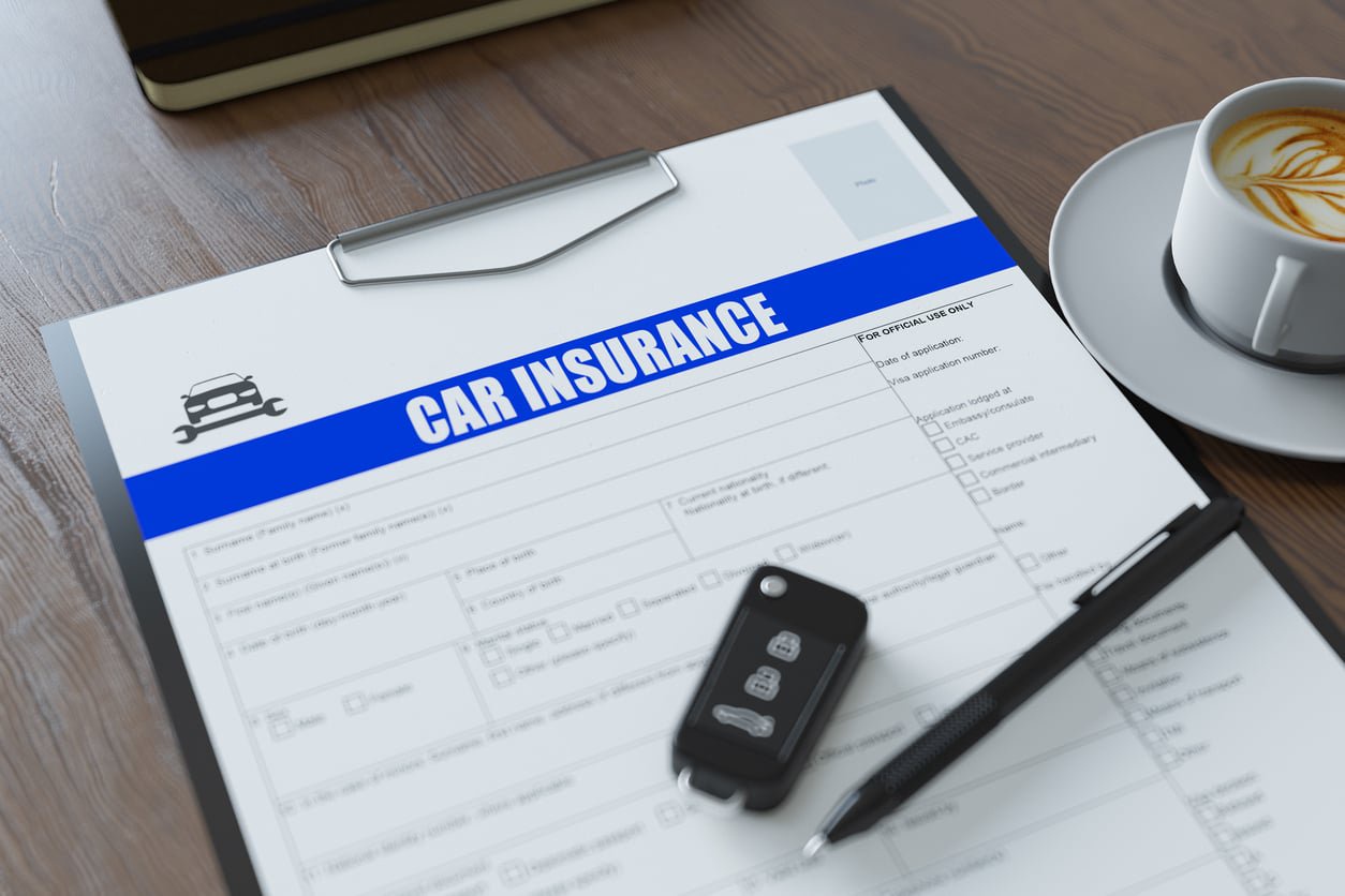 A car insurance form on a clipboard sits on a wooden table. Next to it are car keys, a pen, and a cup of coffee with latte art.