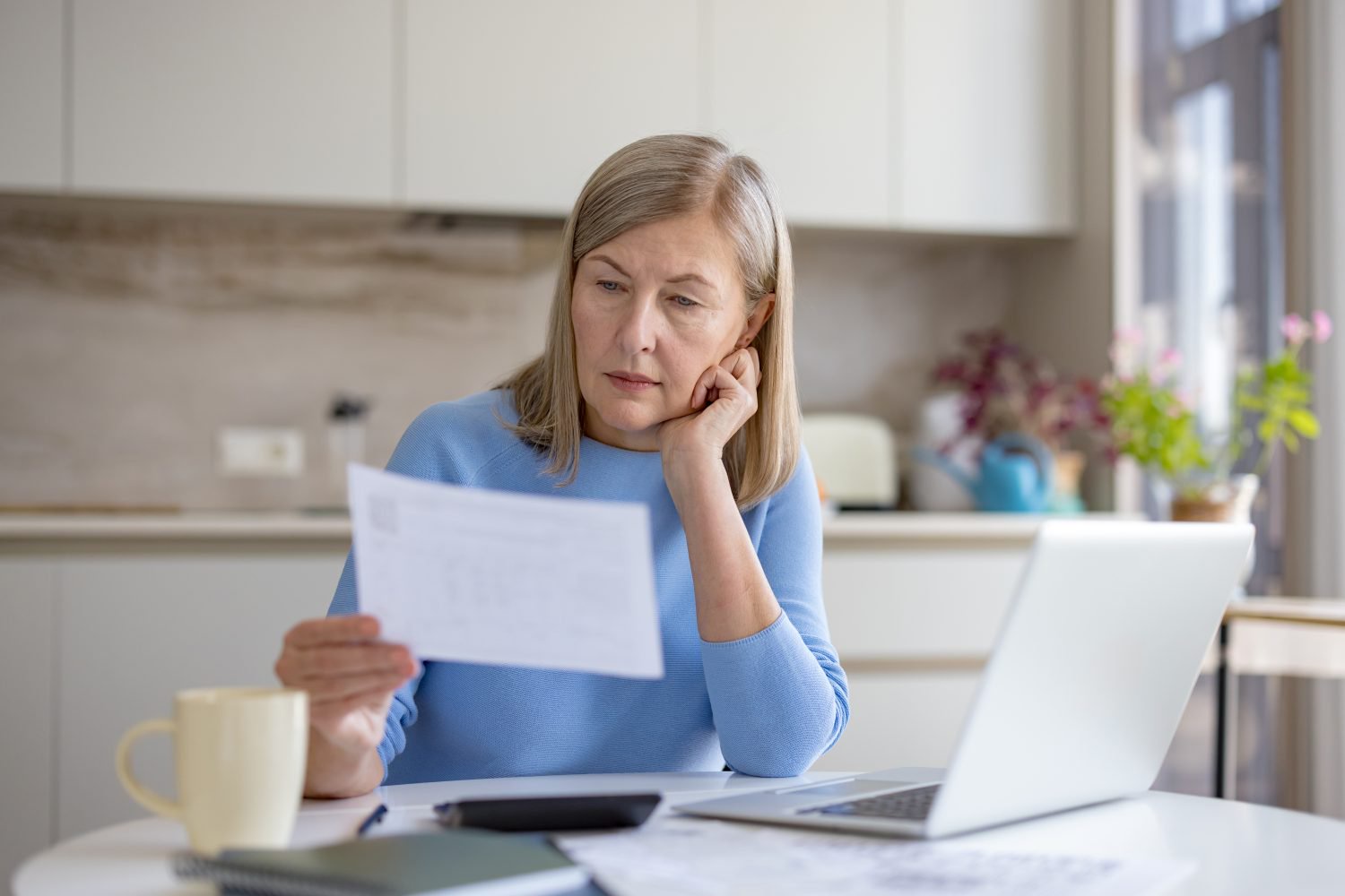 A middle-aged woman with light brown hair and a light blue long-sleeved shirt sits at a white table, holding a white document in her hands and looking at it with a thoughtful expression. To her right, an open silver laptop is visible, and to her left, a light yellow mug and a dark-covered notebook are on the table. The background shows a clean, modern kitchen with light-colored cabinets and a window.