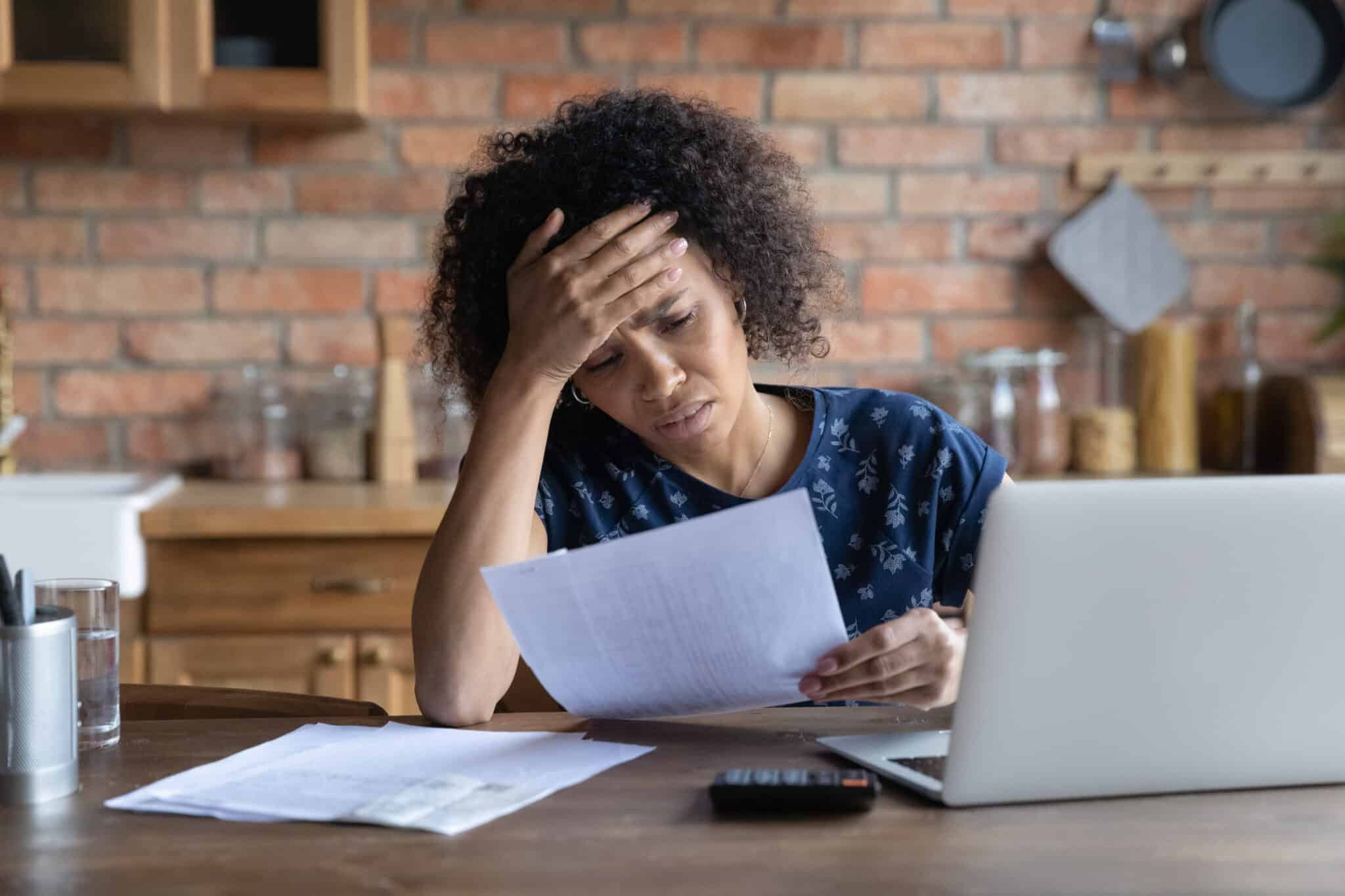A woman with dark curly hair sits at a wooden table, holding a document and looking distressed, with her hand on her forehead. A laptop, calculator, glass of water, and pen holder are on the table. The background shows a brick wall kitchen with wooden cabinets.