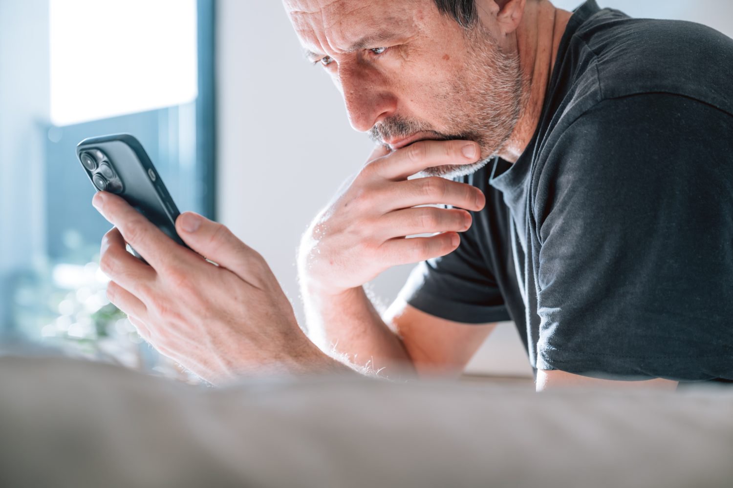 Authentic middle-aged caucasian man looking extremely worried while staring at smartphone, hand on chin. Soft natural daylight from window. Powerful concept for bad news, debt, stress, anxiety, crisis