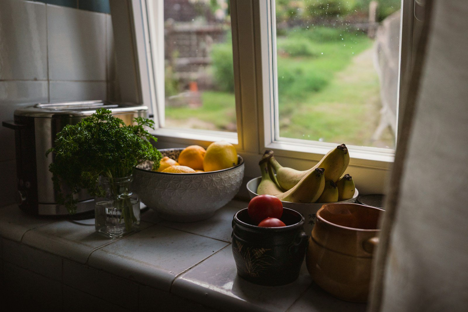 A bowl of lemons, bananas, and tomatoes on a kitchen windowsill with parsley
