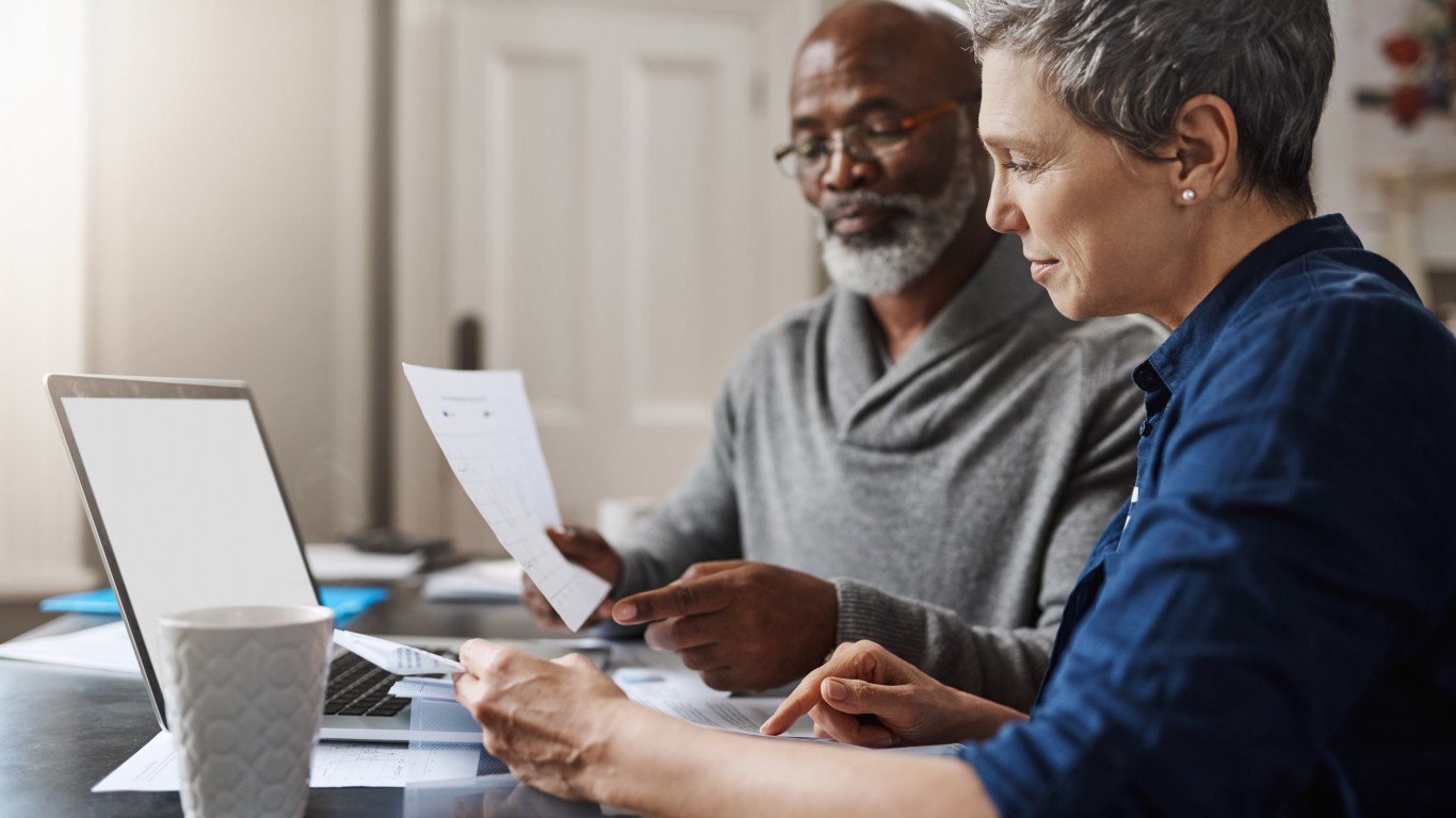 A focused older couple sits at a table, reviewing financial papers and a laptop. The woman, with short gray hair and a blue shirt, points to a document while the man, wearing glasses and a gray shawl-collar sweater, holds another paper and points towards the table. A white mug is in the foreground next to the laptop with a blank screen. The scene conveys a sense of joint financial planning.