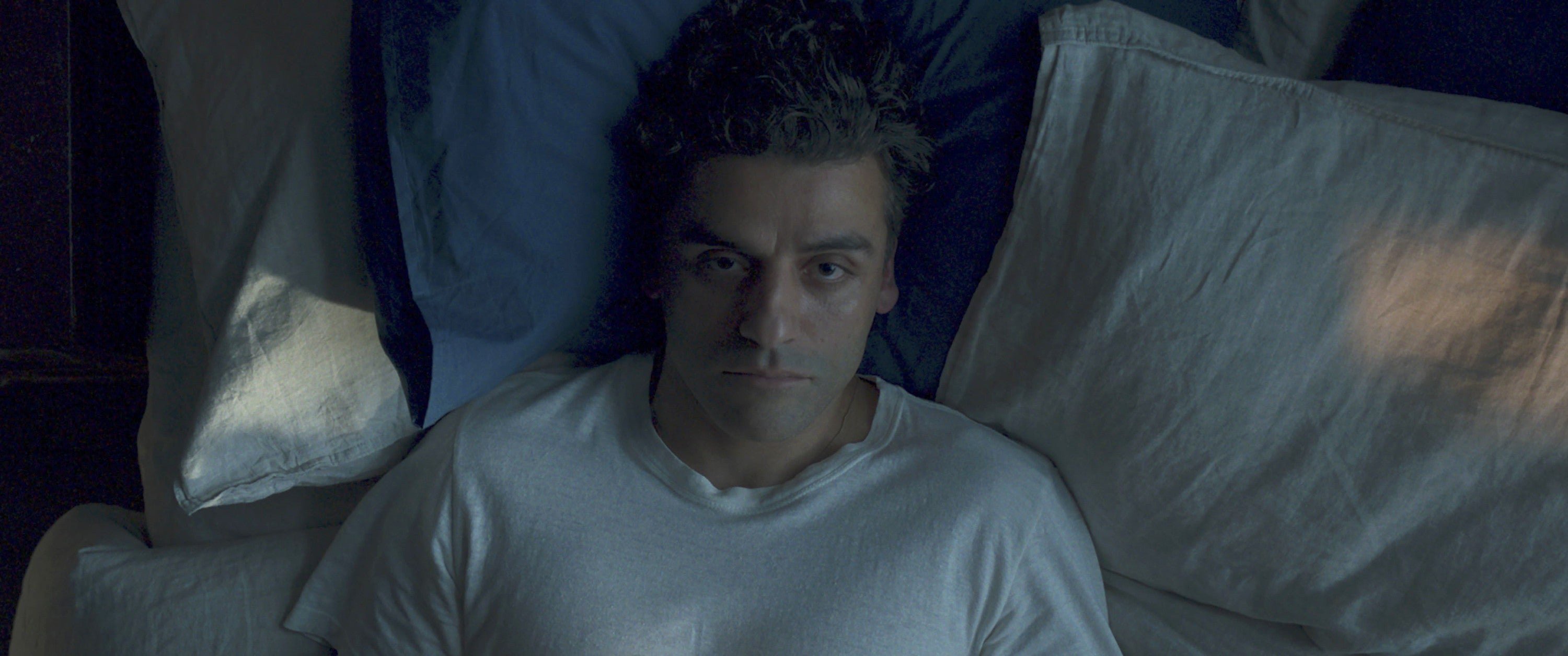A person lies in bed staring upward, wearing a plain T-shirt, conveying a contemplative or pensive mood