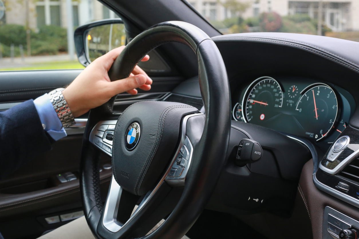 A person wearing a watch is holding the steering wheel of a BMW car. The dashboard shows various gauges and controls, and the view through the windshield reveals a street and buildings.