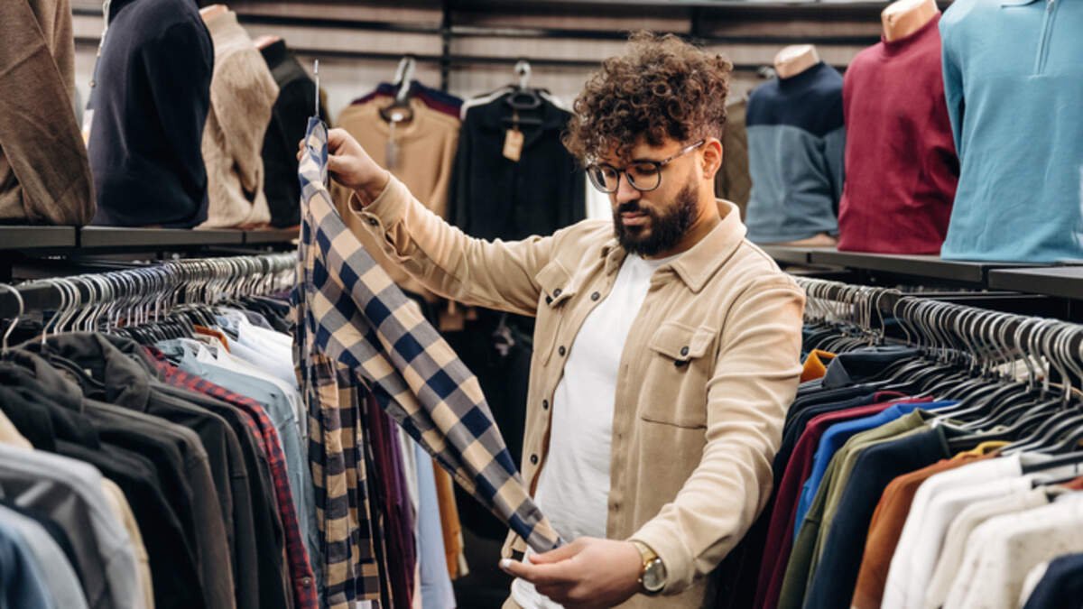 Man with curly hair inspects a plaid shirt while shopping in a clothing store, with a variety of garments displayed on hangers around him
