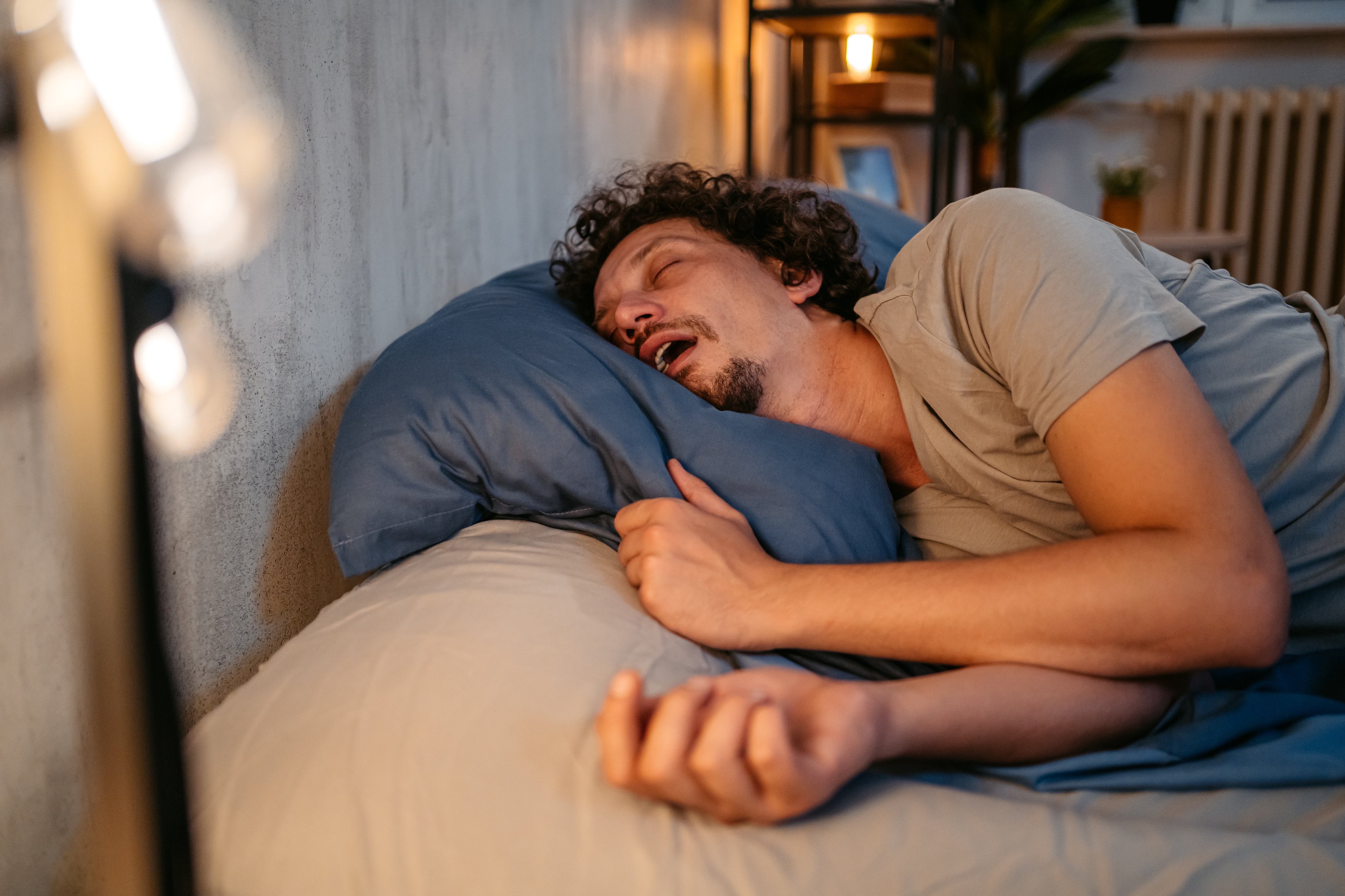 A person asleep on their side in bed, mouth slightly open, with a relaxed expression, in a dimly lit room