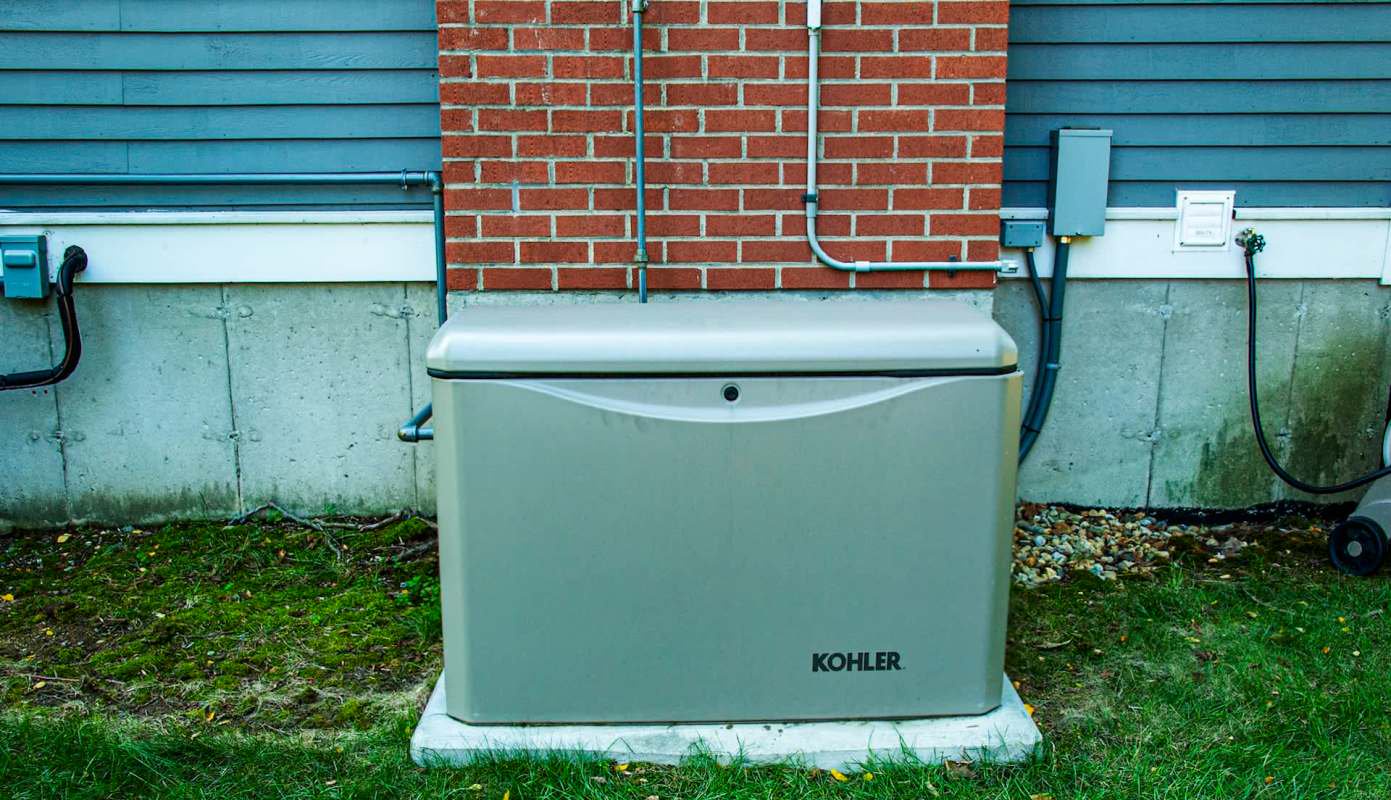 A whole-home generator placed against a brick wall with electrical connections and grass nearby.