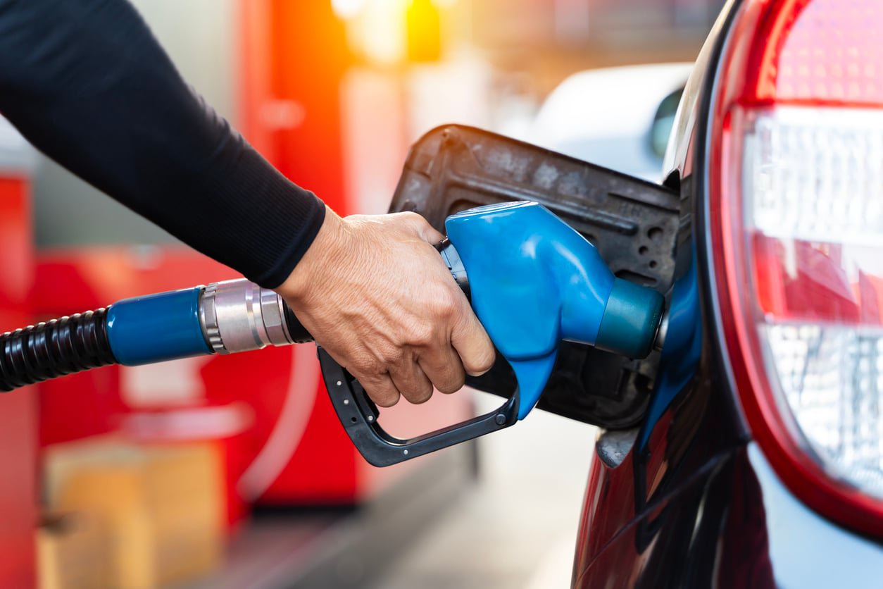 A person refueling a car at a gas station. The individual's hand holds a blue fuel nozzle inserted into the car's fuel tank. The vehicle is dark-colored, and a red gas pump is visible in the background.