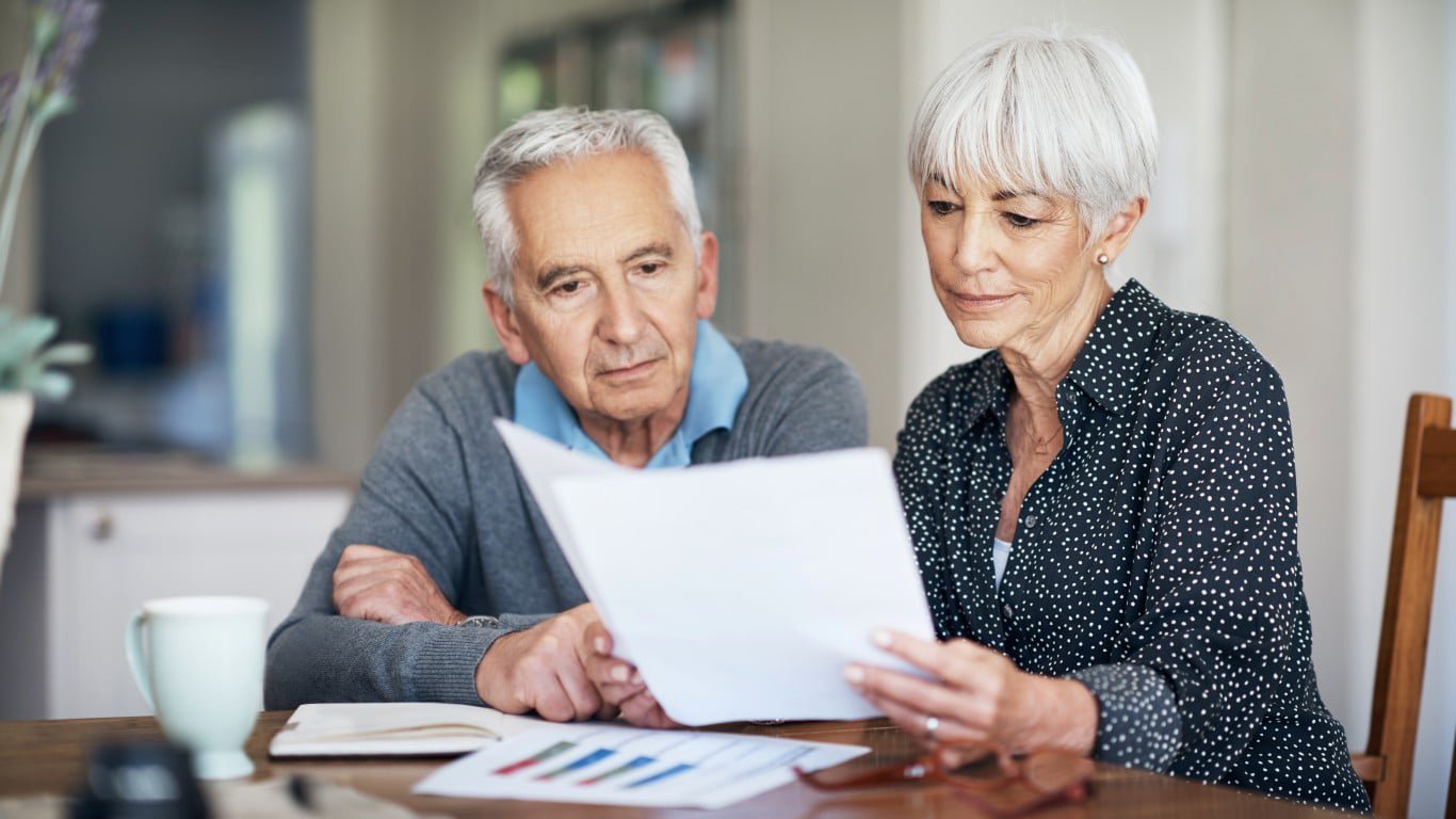 A senior couple sits at a wooden table, with the woman holding and looking at several white papers while the man looks over her shoulder. The woman has short grey hair and wears a black polka-dotted shirt. The man has grey hair and wears a blue collared shirt with a grey sweater. On the table are a light blue mug, an open notebook, and printed documents featuring colorful bar graphs. The background shows a softly blurred home interior.