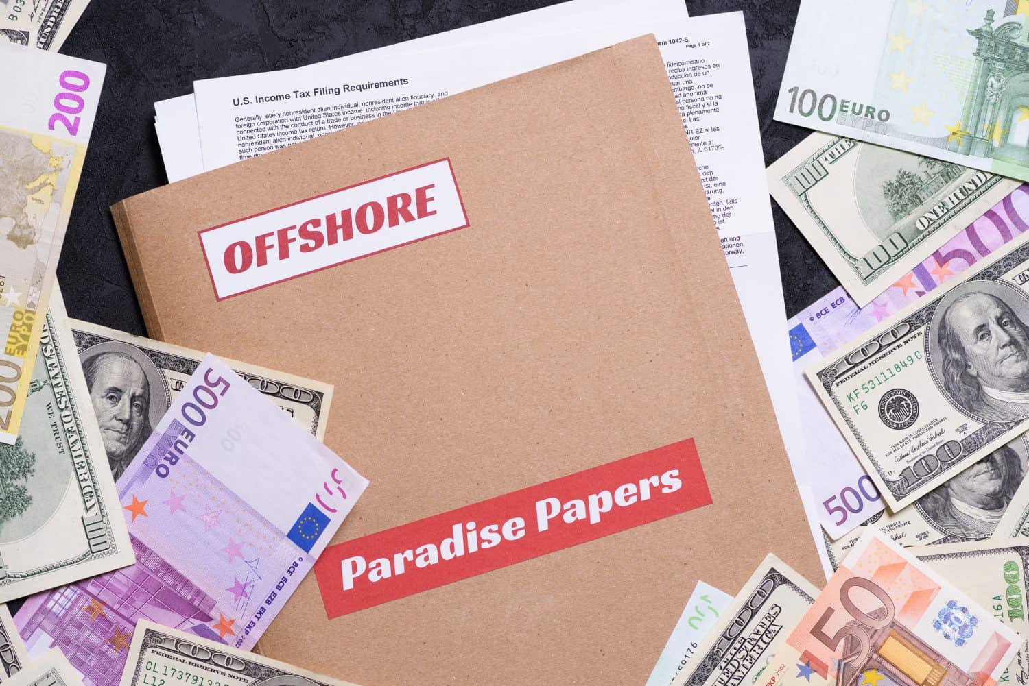 A flat lay composition shows a light brown folder with two red labels: "OFFSHORE" at the top and "Paradise Papers" at the bottom. Surrounding the folder are scattered banknotes of various denominations, including US dollars (100 and 50 notes) and Euros (50, 100, 200, and 500 notes). A white document titled "U.S. Income Tax Filing Requirements" is partially visible beneath the folder, all set against a dark, textured background.