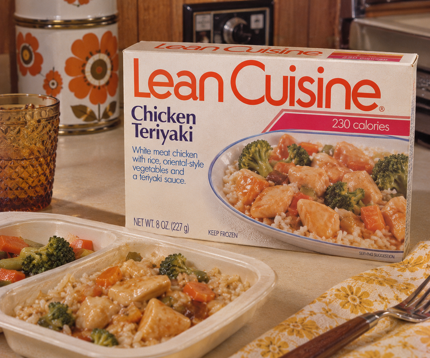 A Lean Cuisine Chicken Teriyaki frozen meal sits on a table beside its box, with rice, vegetables, and chicken visible. A drink and napkin are nearby. The meal and packaging are clearly displayed.