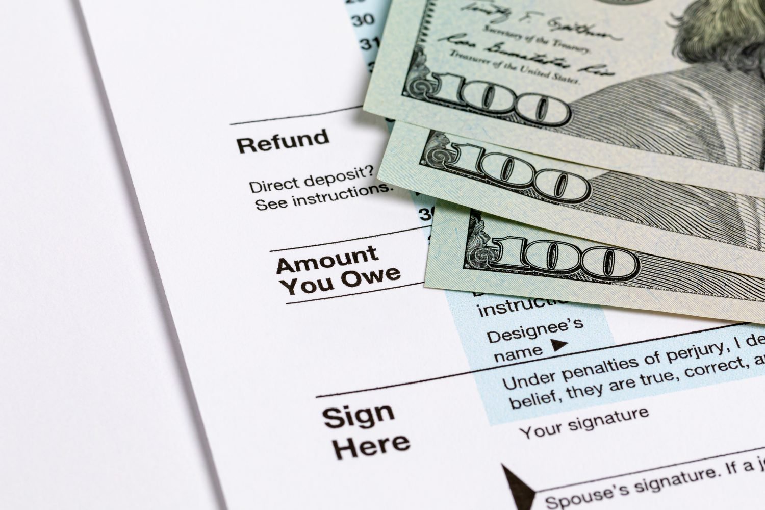 Three US one-hundred-dollar bills are fanned out and partially cover a white tax form. Visible sections of the form include 'Refund', 'Amount You Owe', 'Direct deposit?', and 'Sign Here'. The bills feature a portion of Benjamin Franklin's portrait and the '100' denomination, set against a light background.