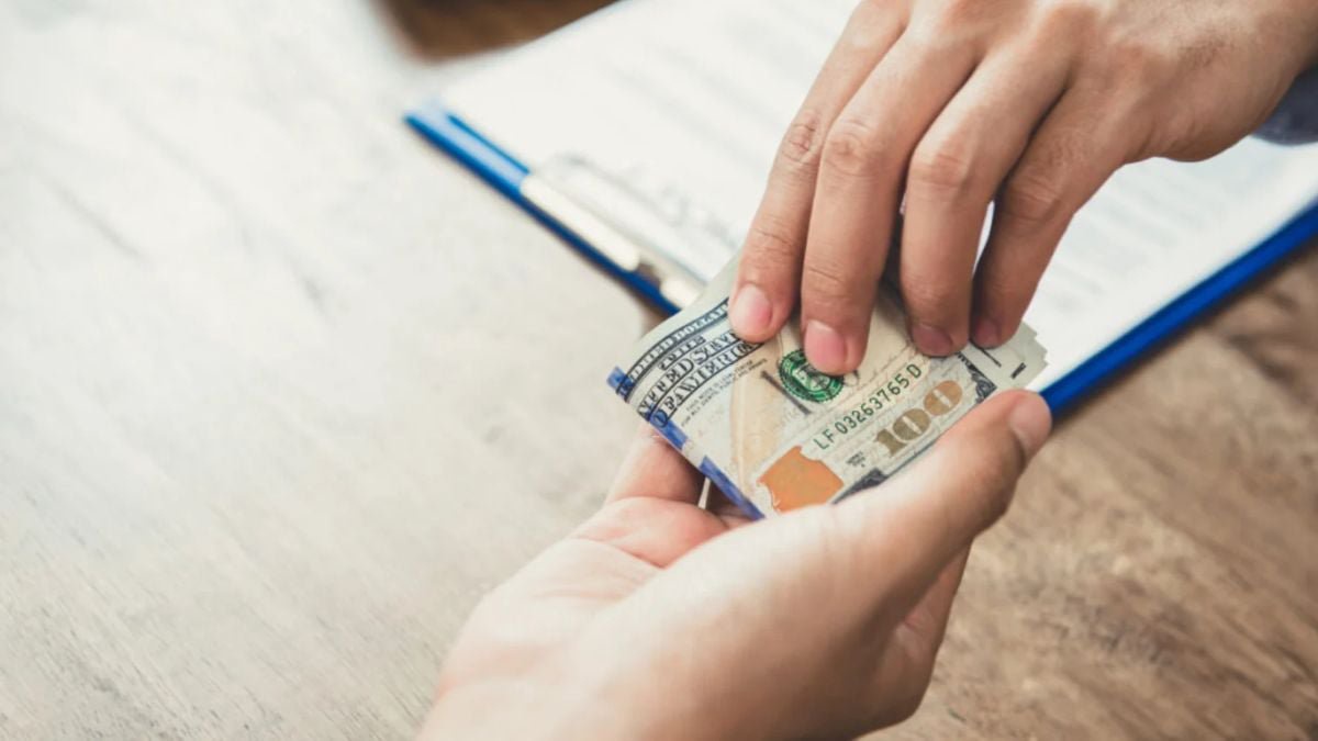 One person handing a stack of US hundred-dollar bills to another person over a clipboard.