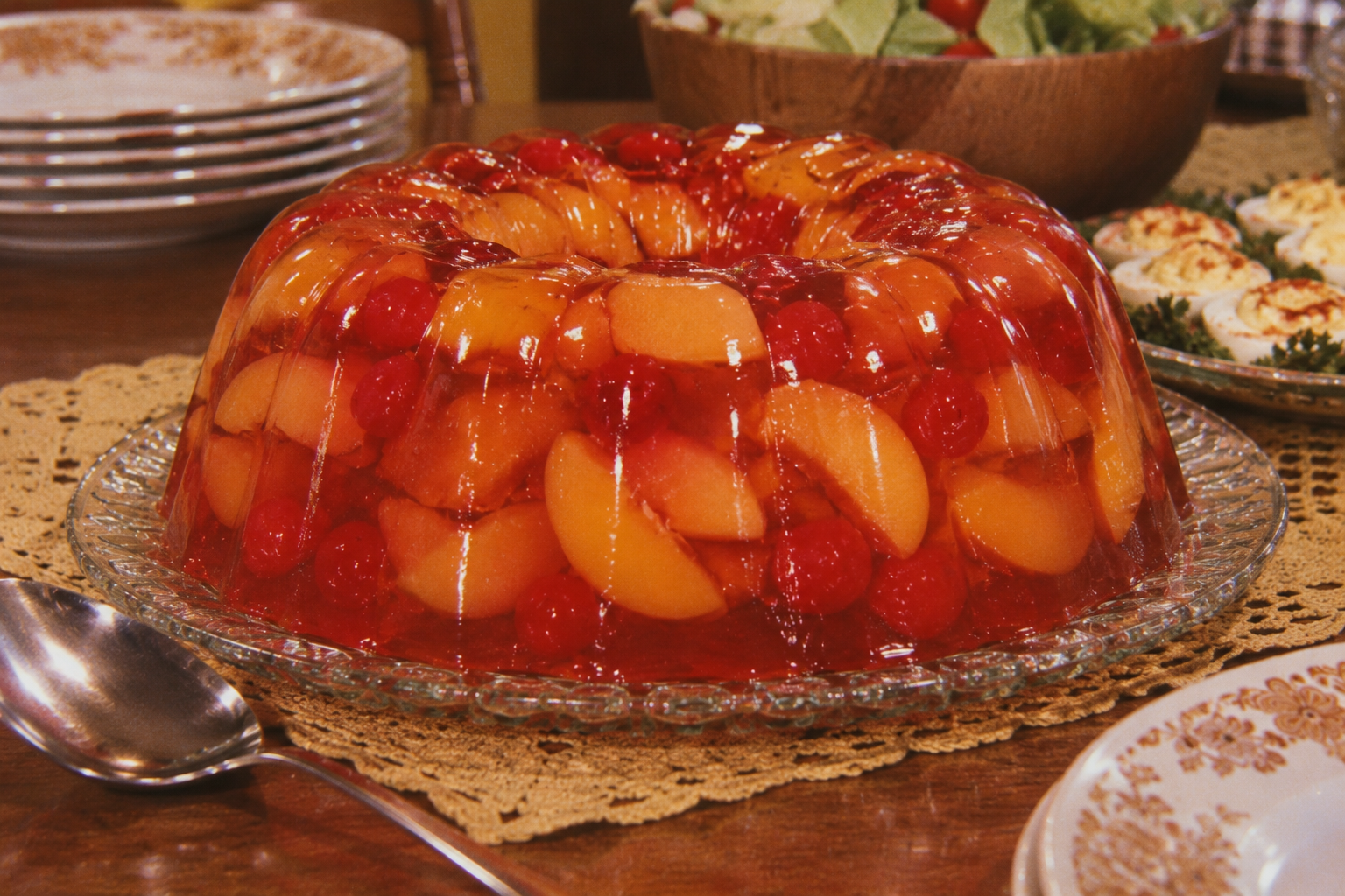 A gelatin mold with peach slices and cherries sits on a glass platter, surrounded by plates, a salad bowl, deviled eggs, and a serving spoon on a lace tablecloth.