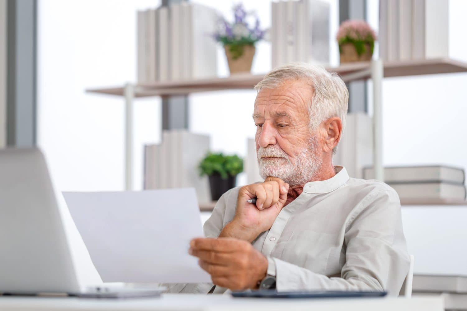 A gray-haired, bearded older man in a light-colored collared shirt sits at a desk, looking down at a white sheet of paper he holds in his left hand. His right hand holds a pen and rests under his chin, indicating thoughtfulness. A silver laptop is visible on the desk to his left, and blurred bookshelves with books and potted plants are in the background.