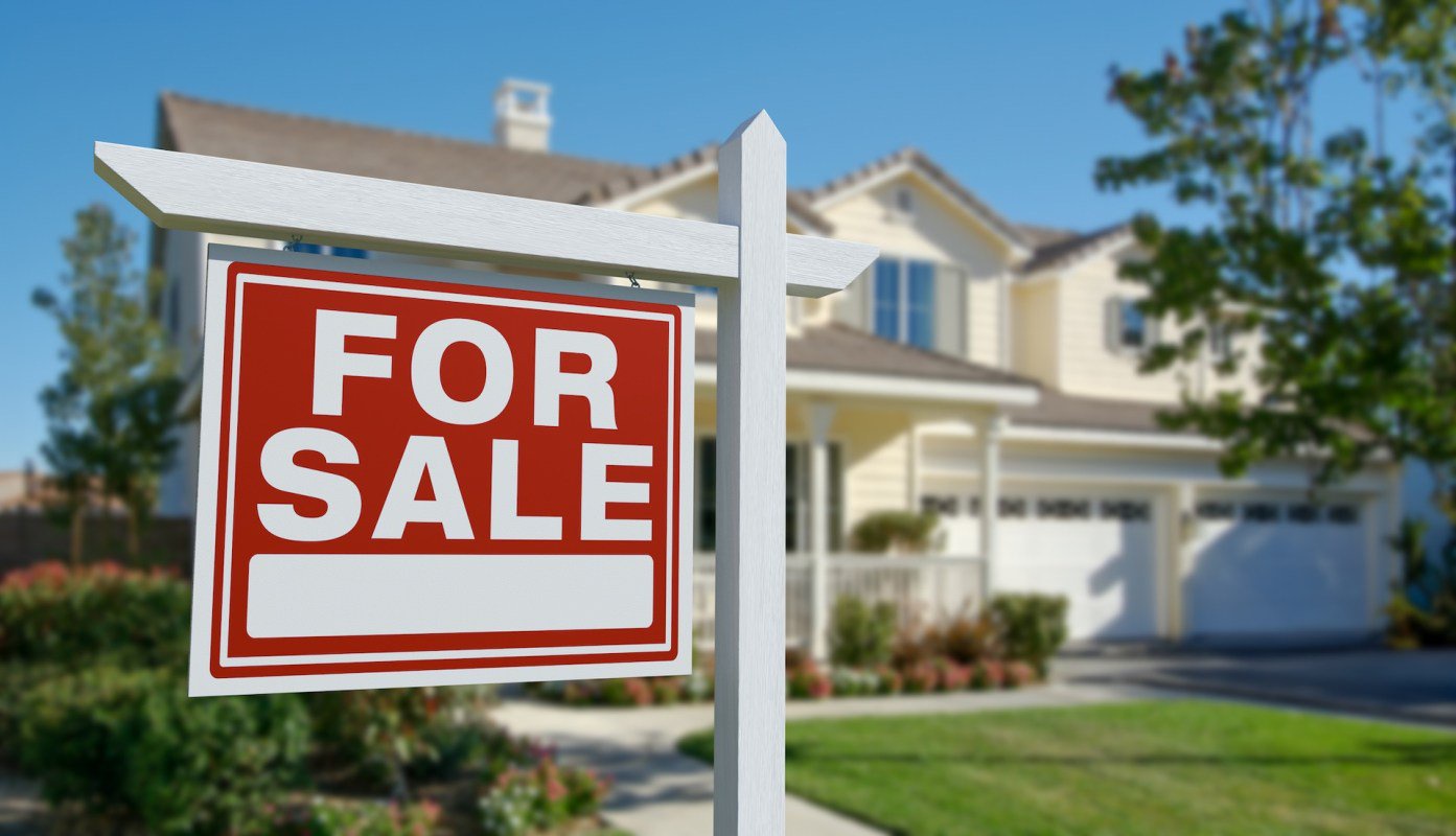 A "For Sale" sign in front of a house with a green lawn and landscaped garden.