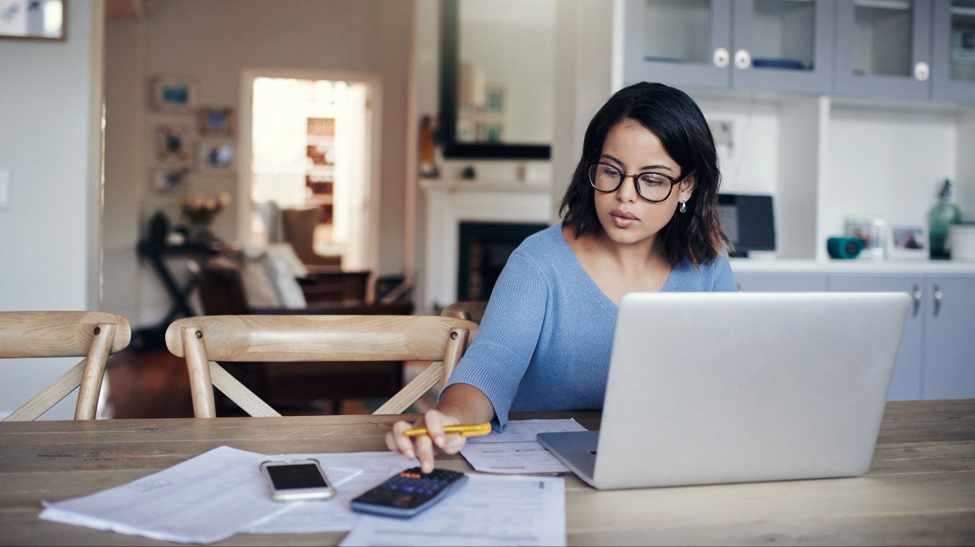 A focused woman with dark hair and glasses, wearing a light blue sweater, sits at a wooden table, intently looking at a silver laptop. Her right hand holds a yellow pencil, positioned near a black calculator and a smartphone, with numerous white financial papers scattered on the table. The soft, blurred background reveals a cozy home interior with a fireplace and kitchen cabinets.