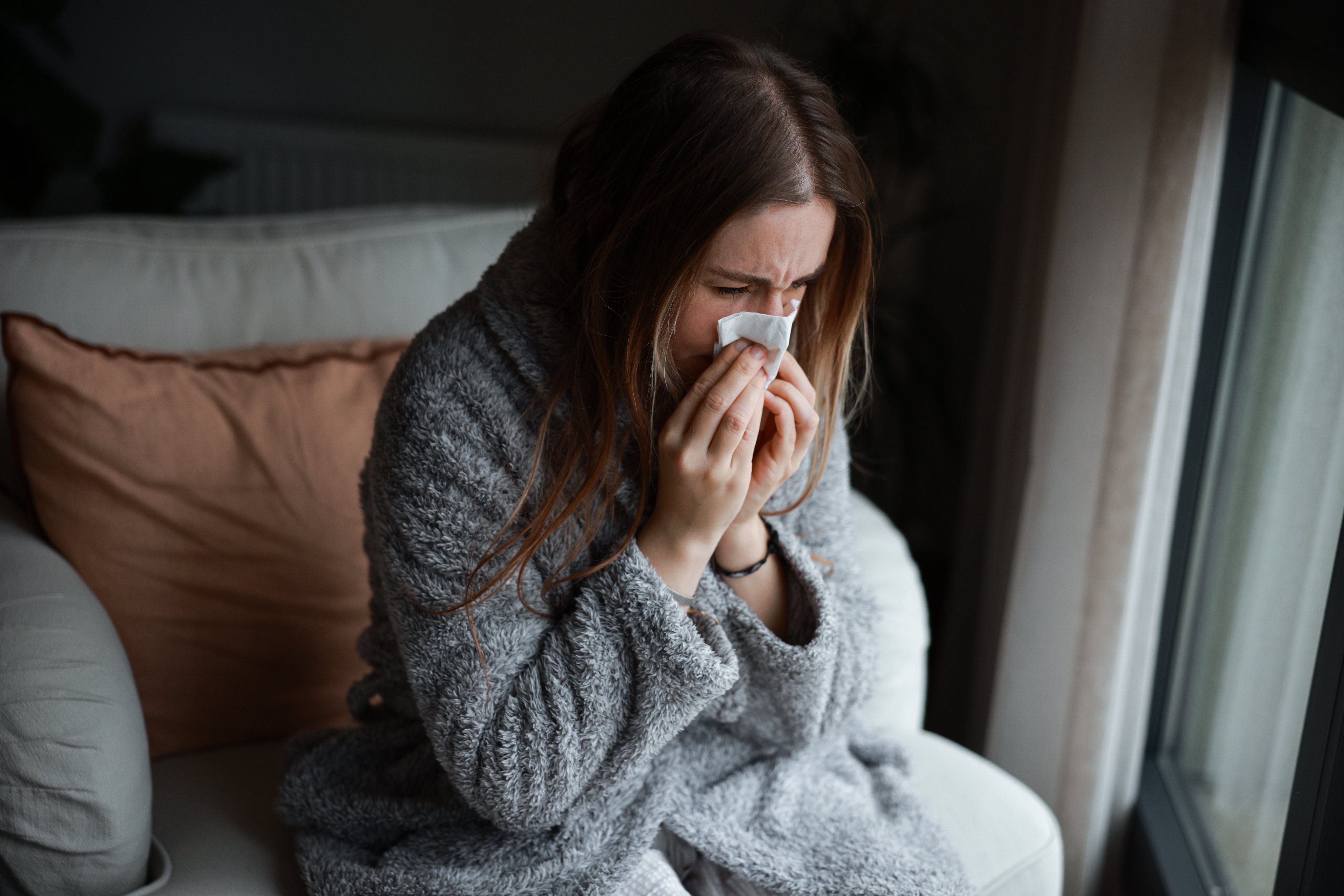 Person in a cozy robe sits on a couch, using a tissue, looking unwell and gazing out a window