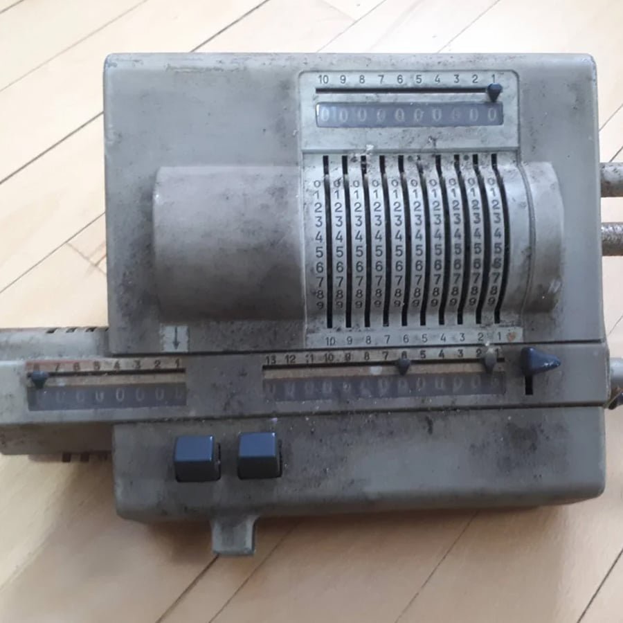 A vintage mechanical calculator with numbered rotating dials and levers sits on a wooden floor. The device appears worn and dusty, showing signs of age and use.