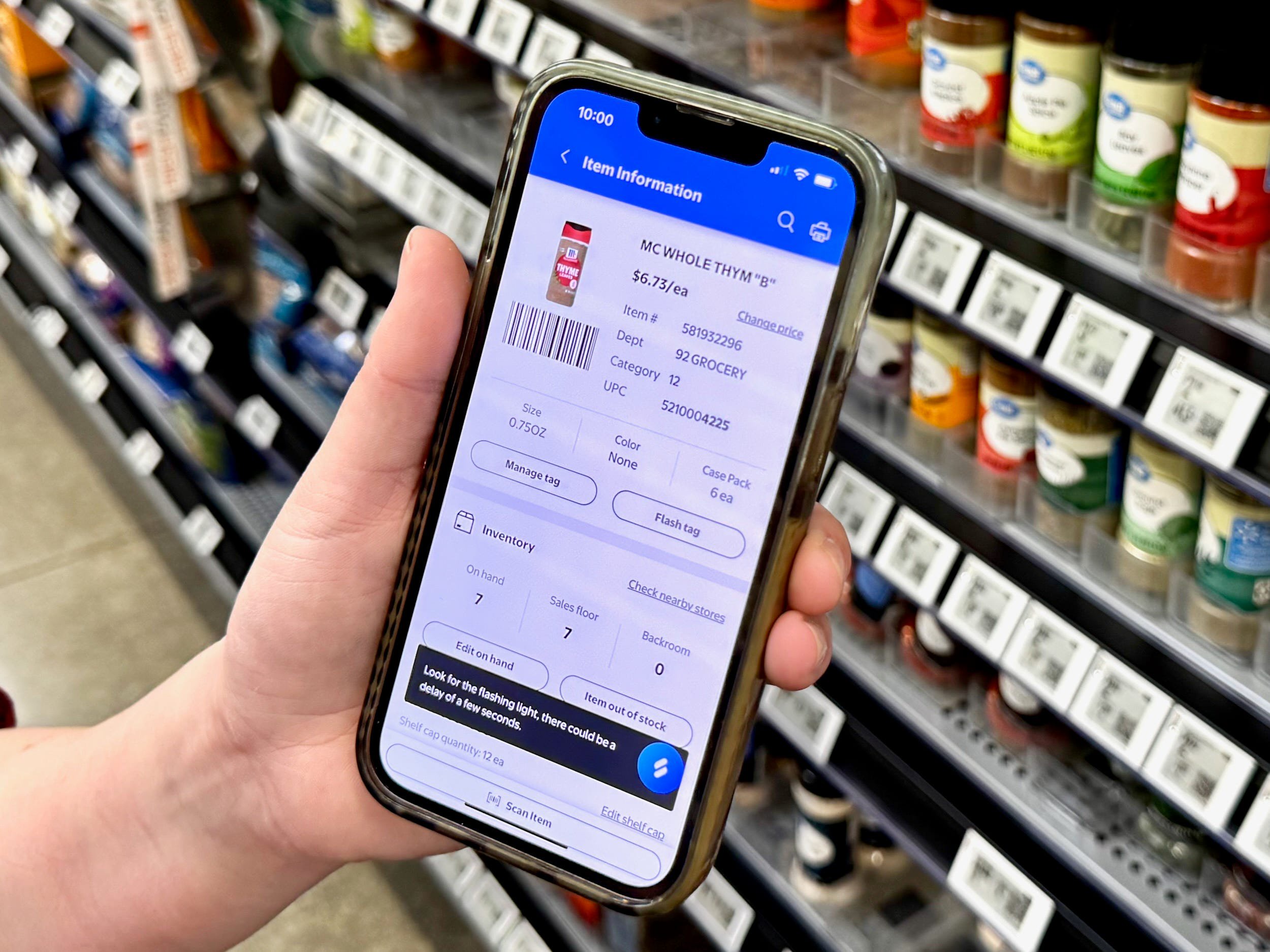 A hand holds a smartphone displaying item information for thyme in front of a spice aisle with digital price tags.