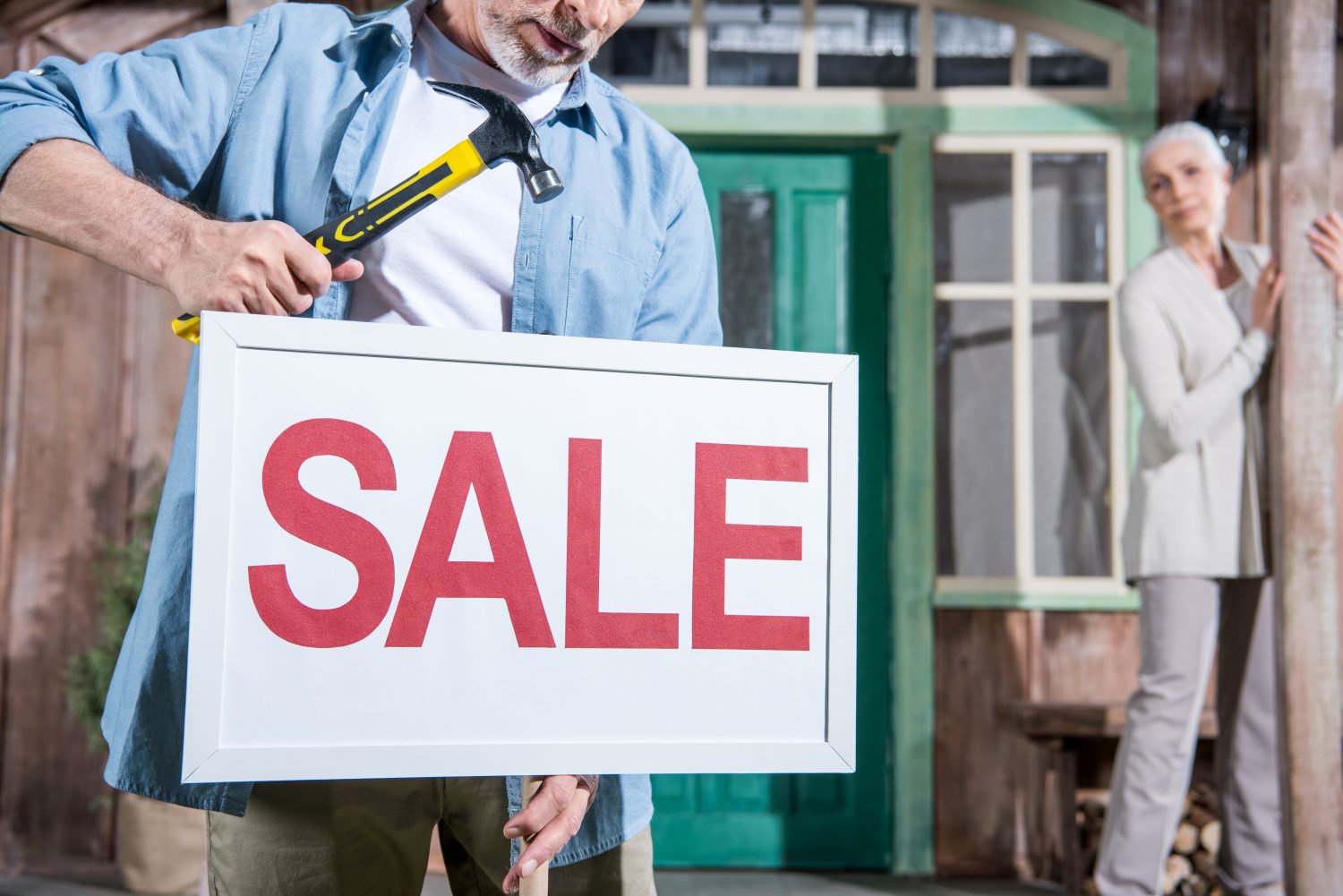 An older man, wearing a blue button-up shirt and khaki pants, holds a white framed 'SALE' sign with prominent red letters and a yellow and black hammer. He stands in front of a rustic wooden house with a green door. An older woman with white hair, wearing a cream cardigan and light pants, is blurred in the background to the right, peering from behind a wooden post.