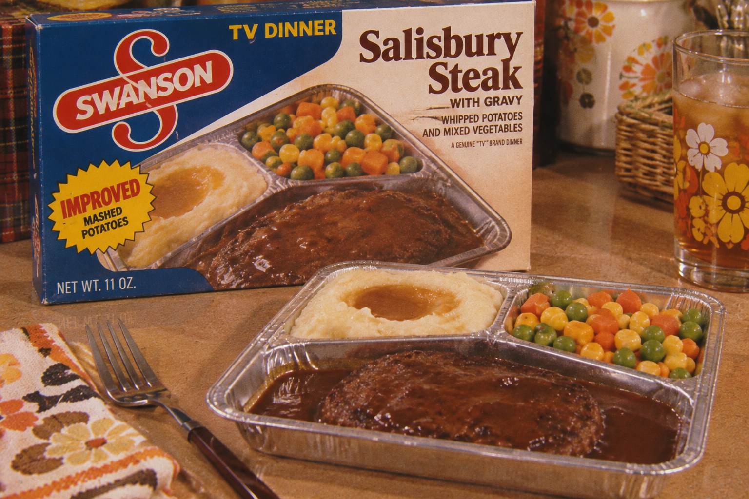 A TV dinner tray with Salisbury steak, mashed potatoes and gravy, and mixed vegetables sits on a table next to its Swanson box, a glass of juice, and a fork on a floral napkin.