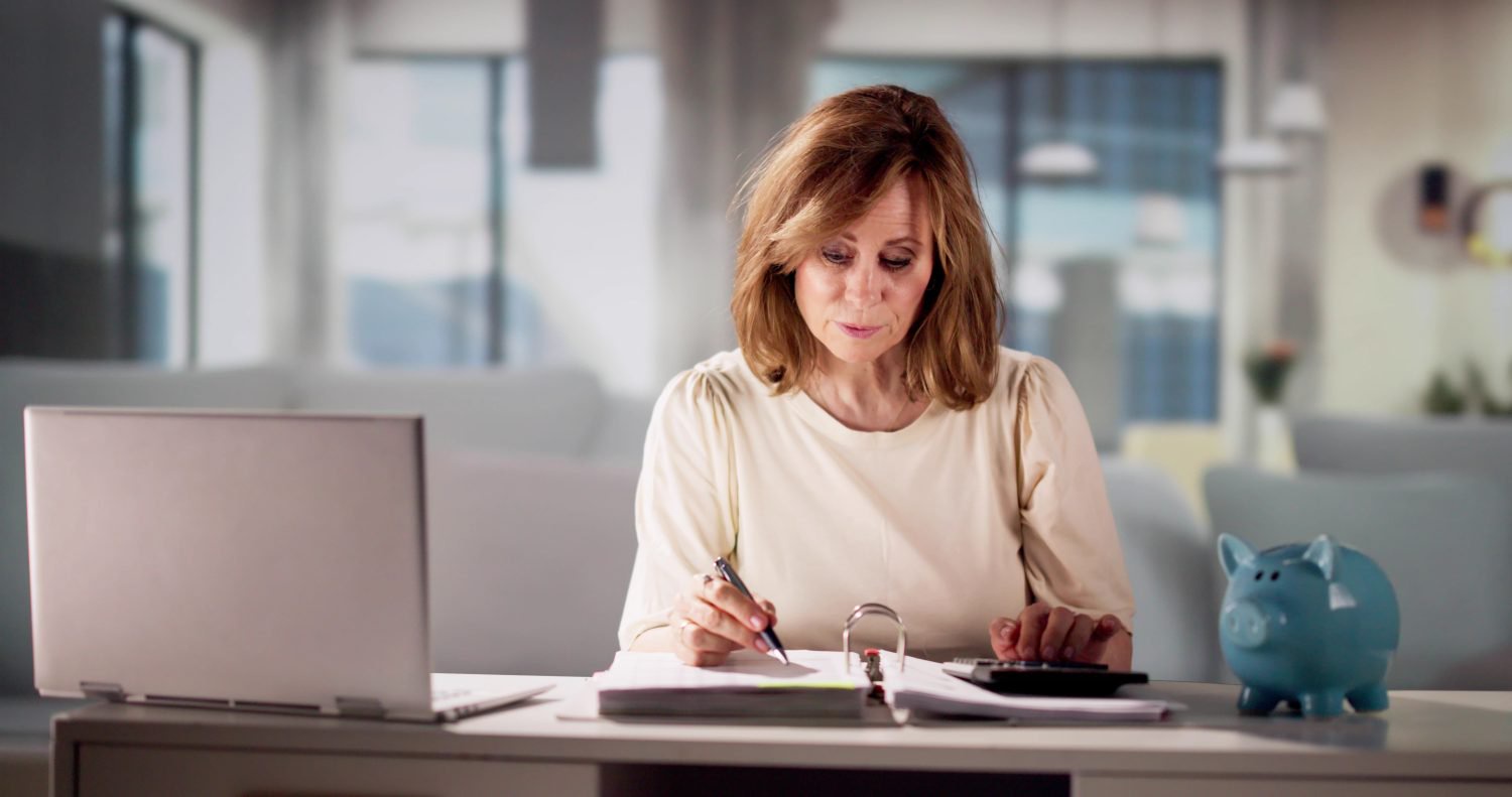 A middle-aged woman with light brown, layered hair sits at a desk, her gaze fixed downwards on documents she is reviewing. She holds a black pen in her right hand, poised over papers, and her left hand rests on a black calculator. To her left, a silver laptop stands open with its screen turned away, and to her right, a light blue ceramic piggy bank sits prominently on the desk. The background is softly blurred, showing a modern, well-lit interior space.