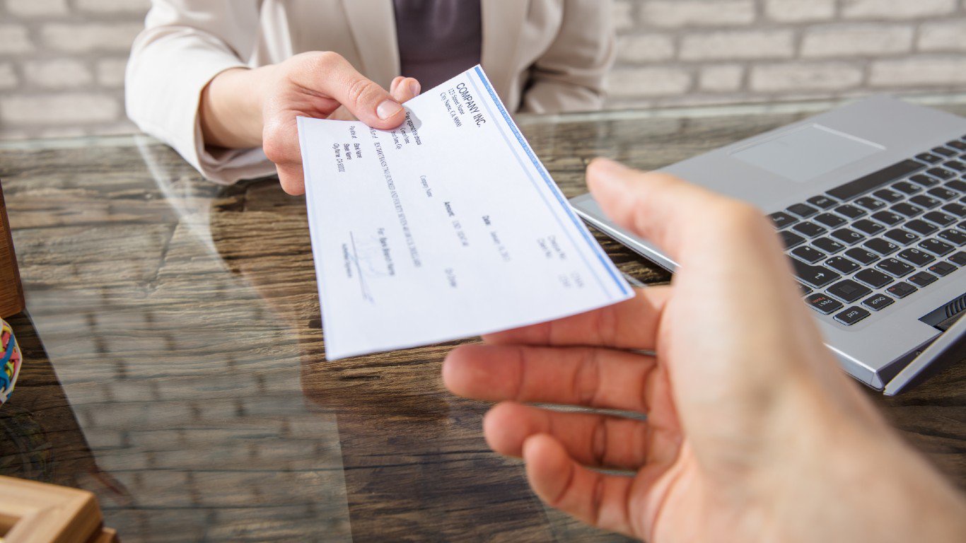 Close-up shot of two people's hands exchanging a white paper check. One hand, belonging to a person in a light-colored jacket, is handing the check to another hand in the foreground. The check has 'COMPANY INC.' printed at the top. A wooden desk surface is visible, along with parts of a silver laptop with a black keyboard on the right side of the frame. A blurry light gray brick wall serves as the background.