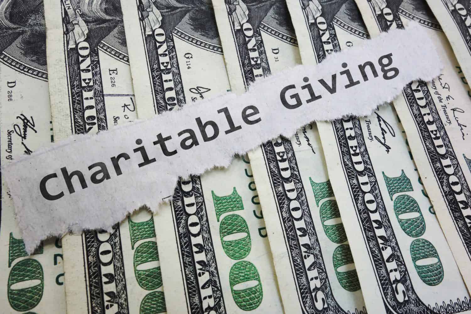 A torn strip of paper with the words 'Charitable Giving' printed in black text lies diagonally across several fanned-out US one hundred dollar bills. The green and white currency shows parts of the '100 DOLLARS' text and signatures.