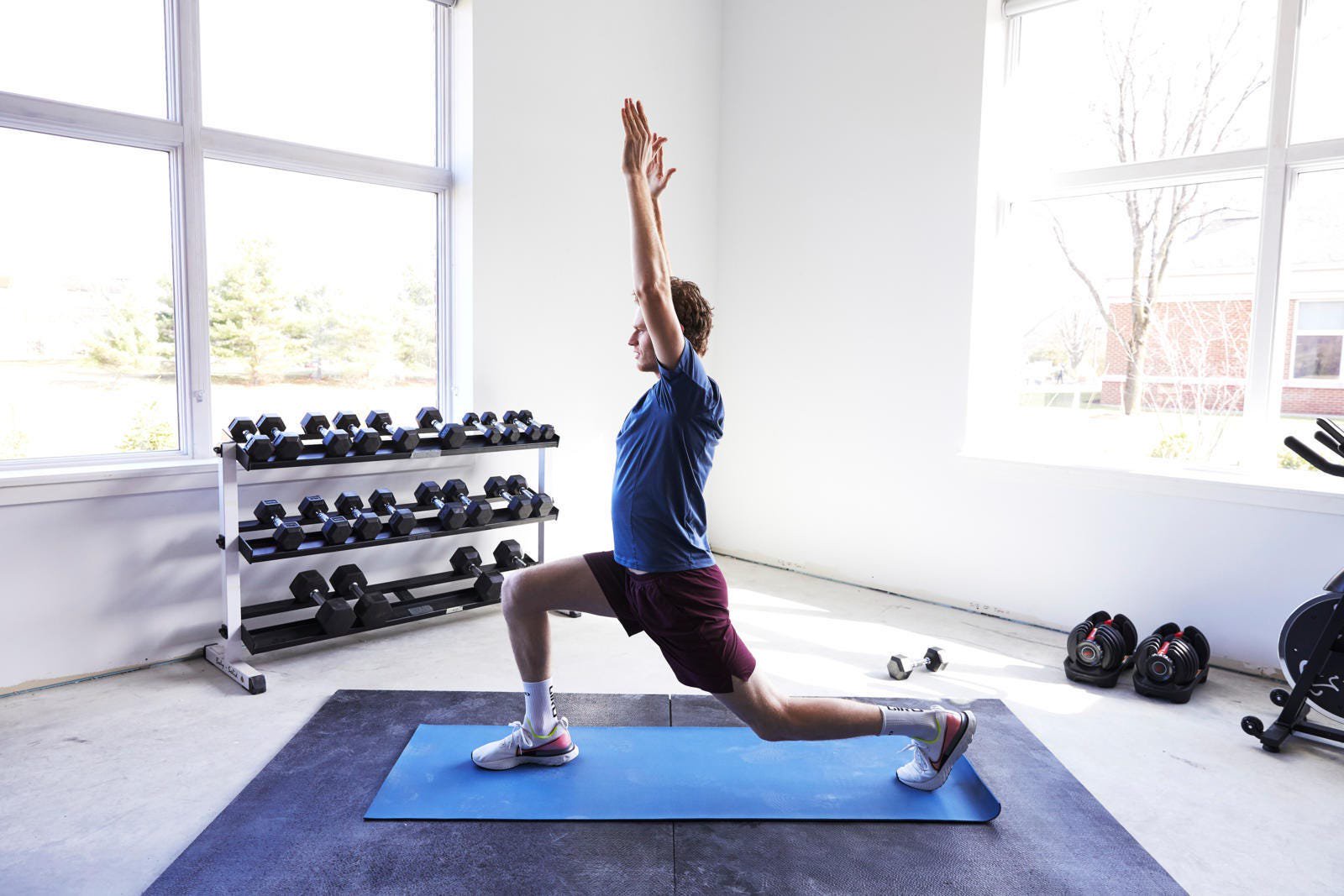 Male exercising in a lunge position with arms raised, in a bright gym with dumbbells in the background.