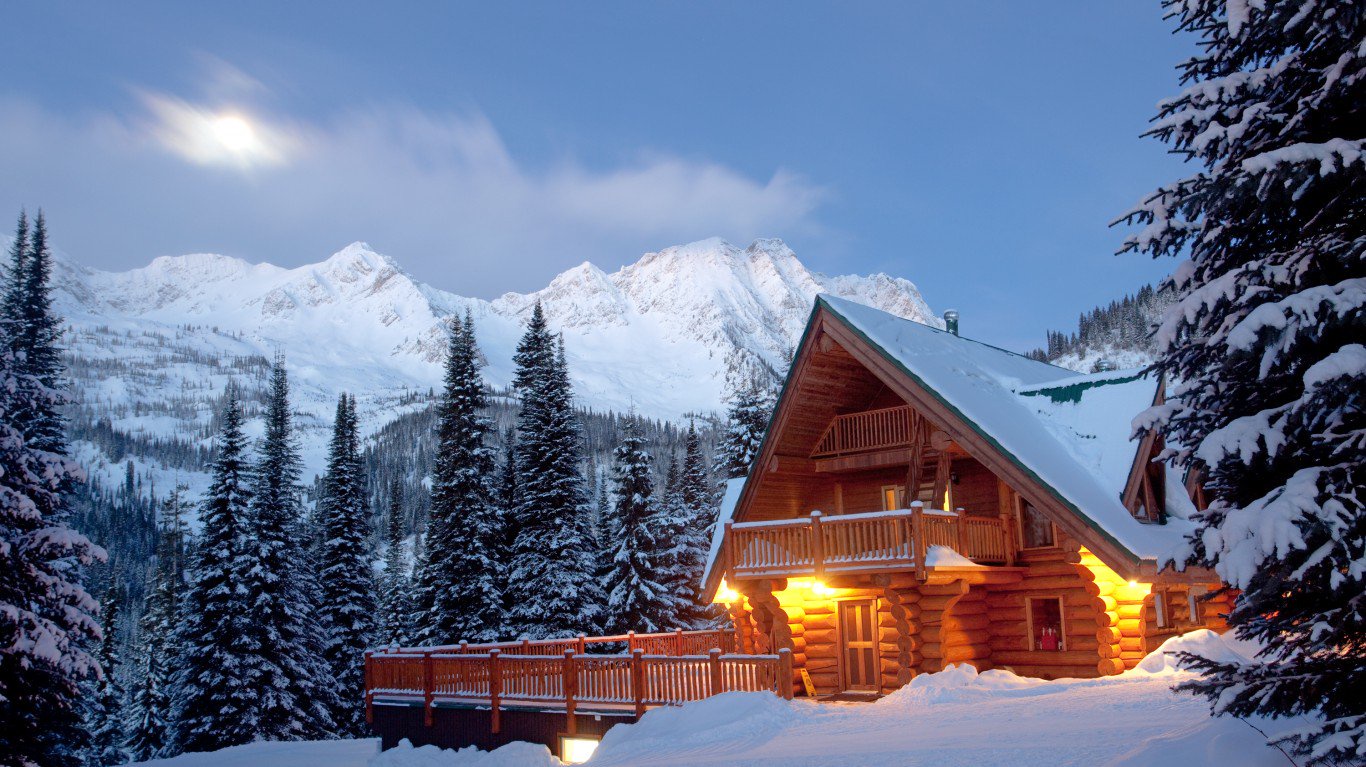 A warm, glowing log cabin stands in a deep snowfield at dusk or dawn. Snow-covered evergreen trees flank the cabin, and majestic snow-capped mountains rise in the background under a blue sky with a bright spot of light, possibly the moon or sun, peeking through clouds. The cabin features a wooden balcony and warm lights emanating from its windows, creating a cozy contrast with the cold winter landscape.