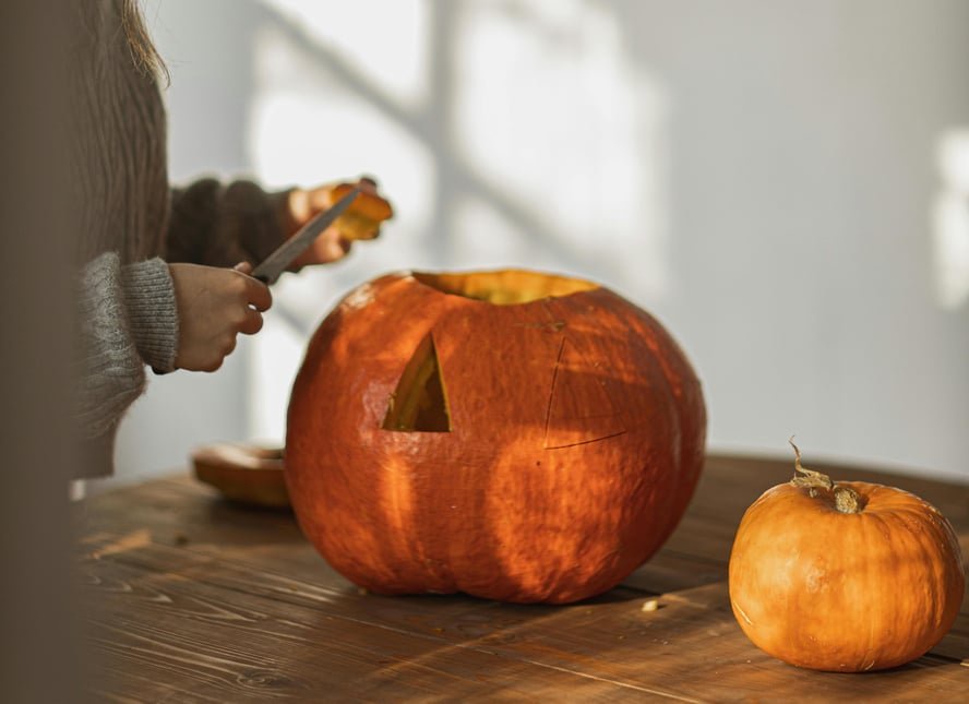 Image of two pumpkins on a table and someone carving a jack-o'-lantern out of one of them.