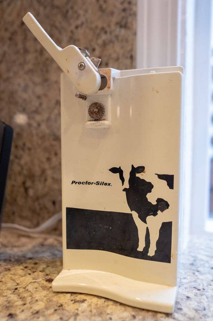A rectangular electric can opener printed with a black and white graphic cow on a black field of grass, with a wavy base foot. 