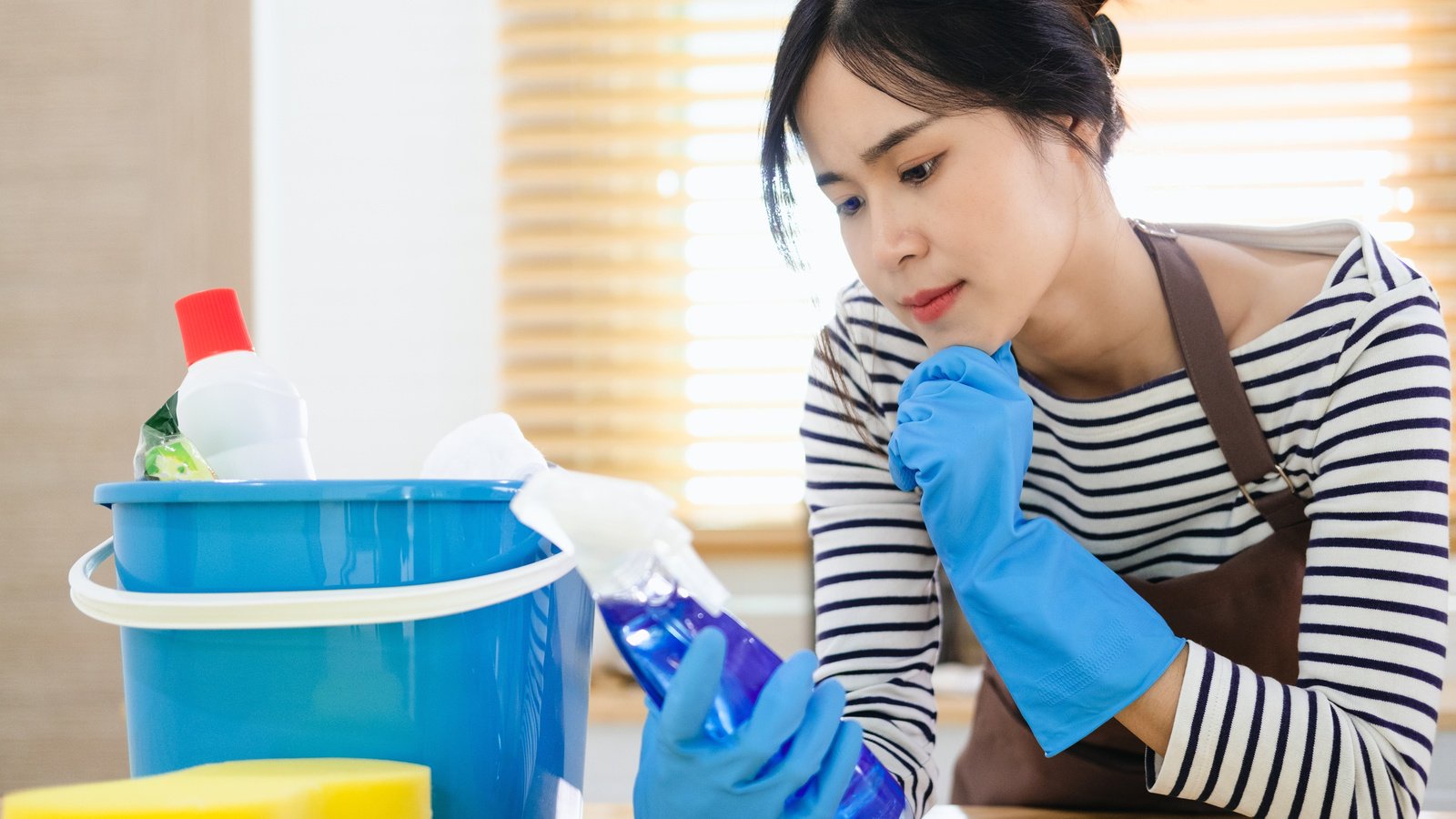 A woman wearing protective gloves examining a cleaning-product bottle beside other cleaning supplies
