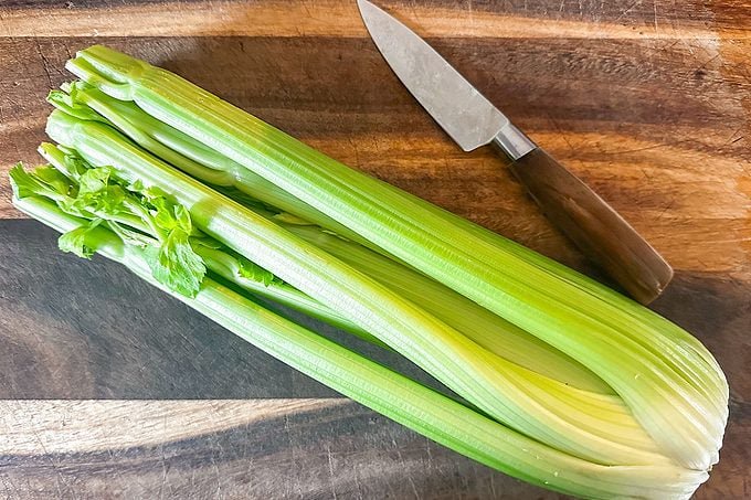 A bunch of fresh celery stalks with leaves sits on a wooden cutting board next to a small knife with a brown handle.
