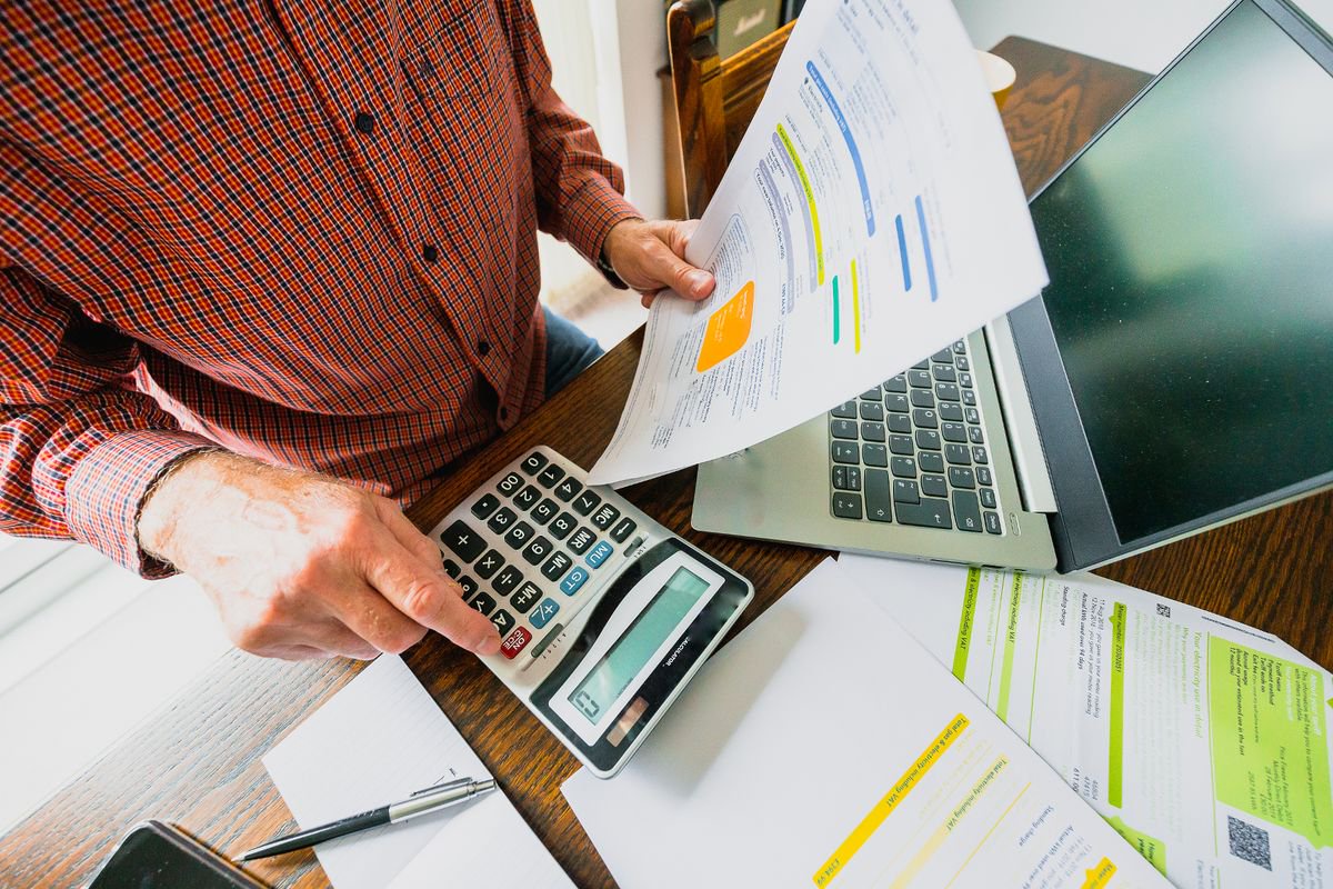 Wide angle image depicting a senior man's hand holding an energy bill while the other hand checks the numbers on a calculator. The table is strewn with documents and also a laptop. Room for copy space.
