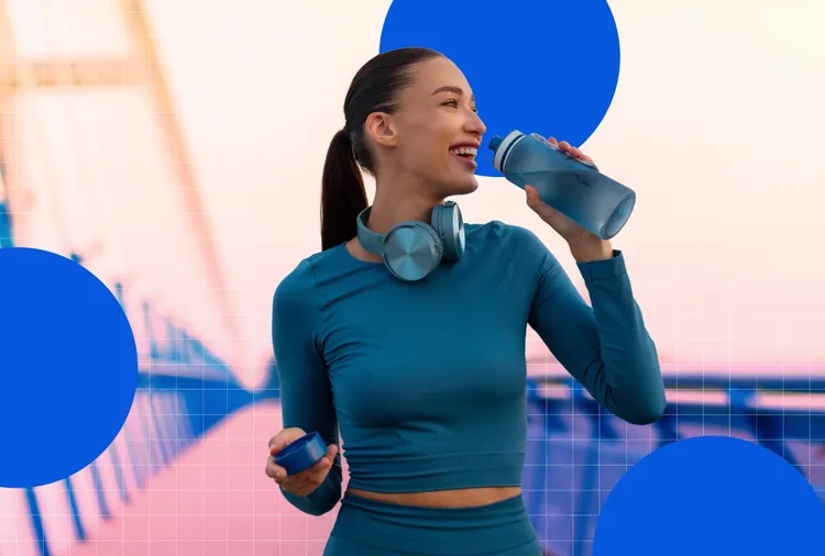 Person wearing workout attire holding a water bottle and standing on a bridge with headphones around their neck