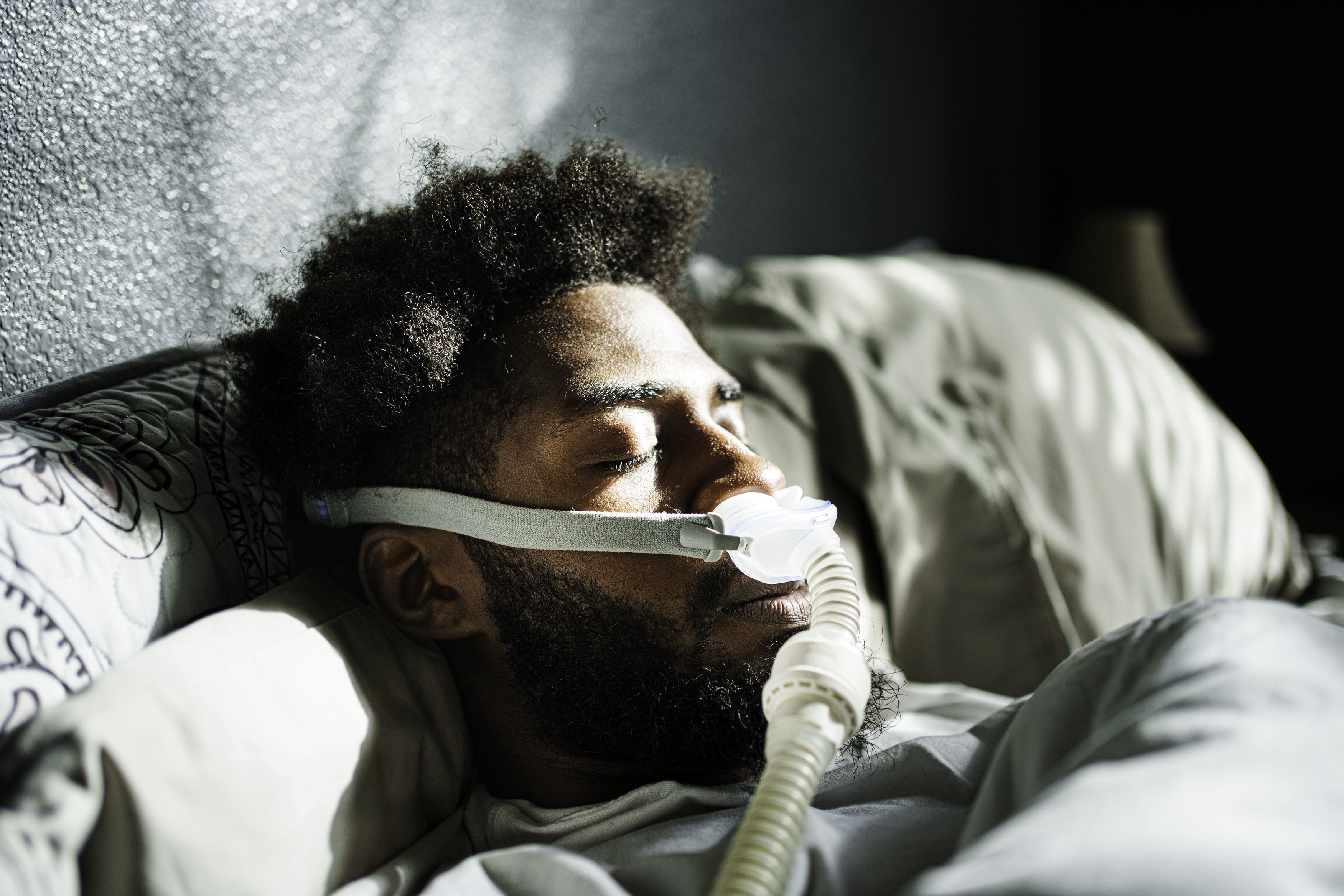 A person sleeping with a CPAP mask on, lying in bed, sunlight illuminating their face