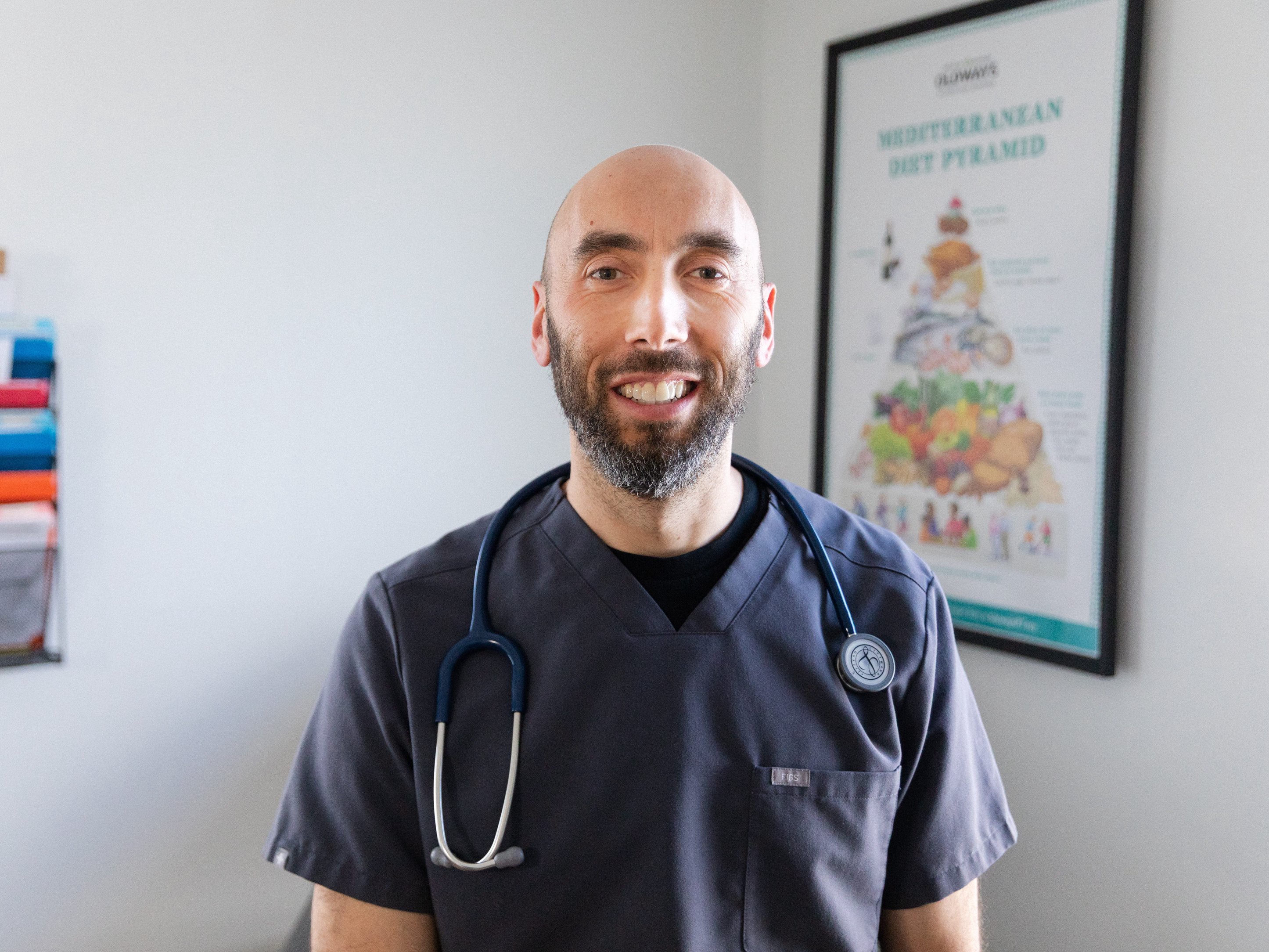 Smiling healthcare worker in dark scrubs with a stethoscope stands in a clinical office with a nutrition poster. Dr_Ben_Hagopian
