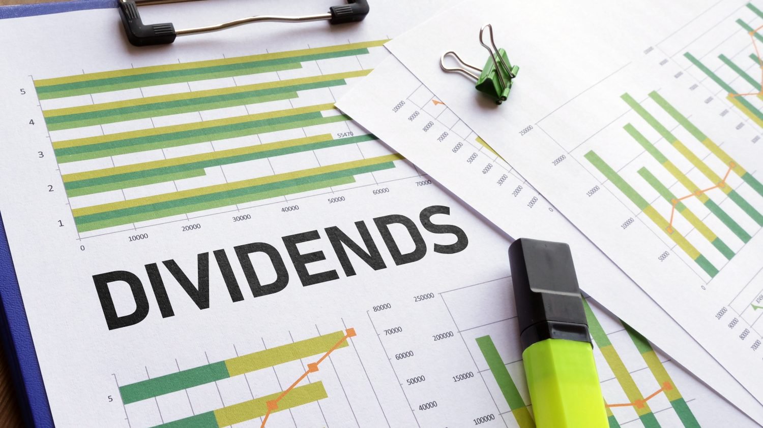 A close-up photograph of multiple financial charts and graphs on white paper, held by a blue clipboard. The word 'DIVIDENDS' is printed in large, bold black letters across the center sheet. A green paperclip holds additional chart papers, and a neon yellow highlighter rests in the foreground. The charts use green and yellow bars along with orange line graphs to illustrate data points.