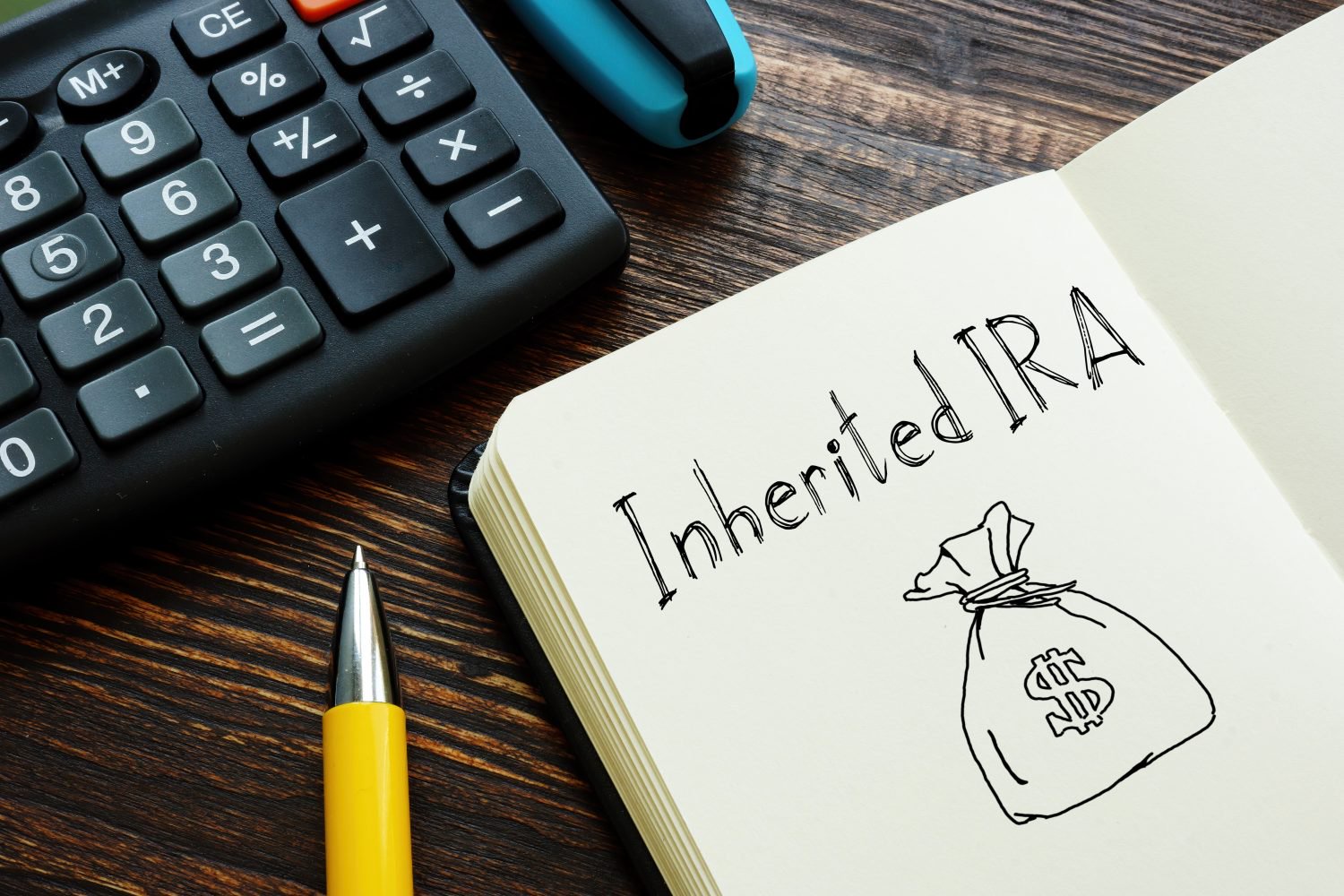 An overhead shot shows a dark wooden desk with a black calculator, a yellow and silver pen, and an open notebook. The notebook's right page displays "Inherited IRA" in black cursive handwriting, below which is a simple line drawing of a money bag with a dollar sign.