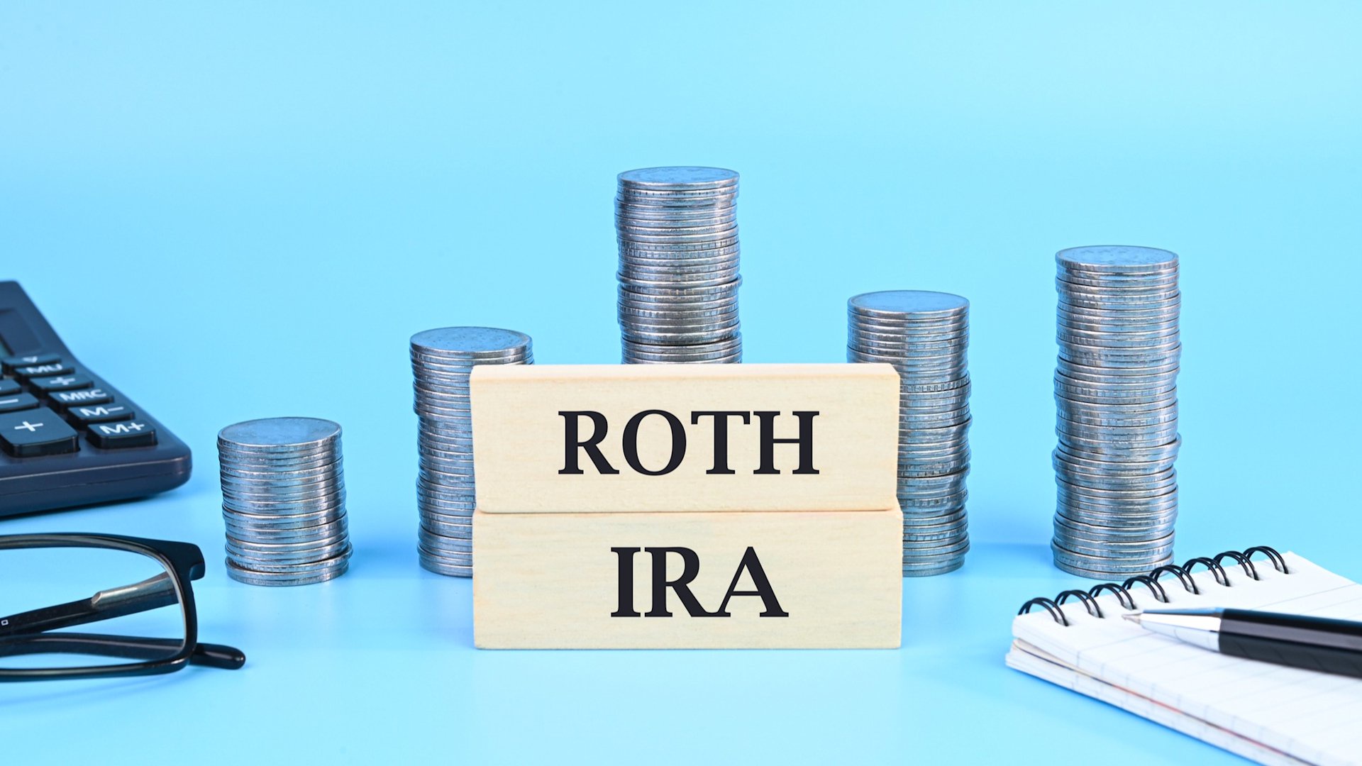 Discover the words Roth IRA on blocks, stacks of coins, a calculator, notepad and pair of glasses