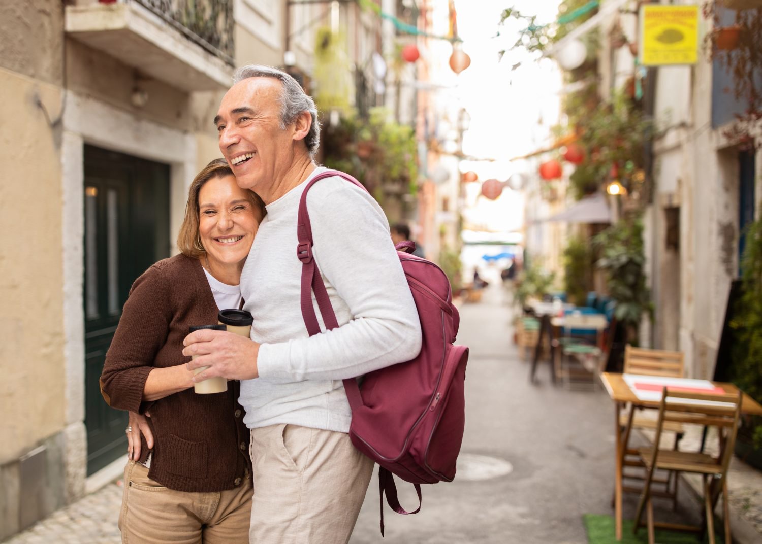 A smiling older man with grey hair, wearing a white sweater and beige pants, with a burgundy backpack, walks with his arm around a smiling older woman. She has brown hair, wears a brown cardigan and beige pants, and holds two takeaway coffee cups. They are on a narrow, sunny European street with stone buildings, hanging decorations, and outdoor cafe seating in the background.