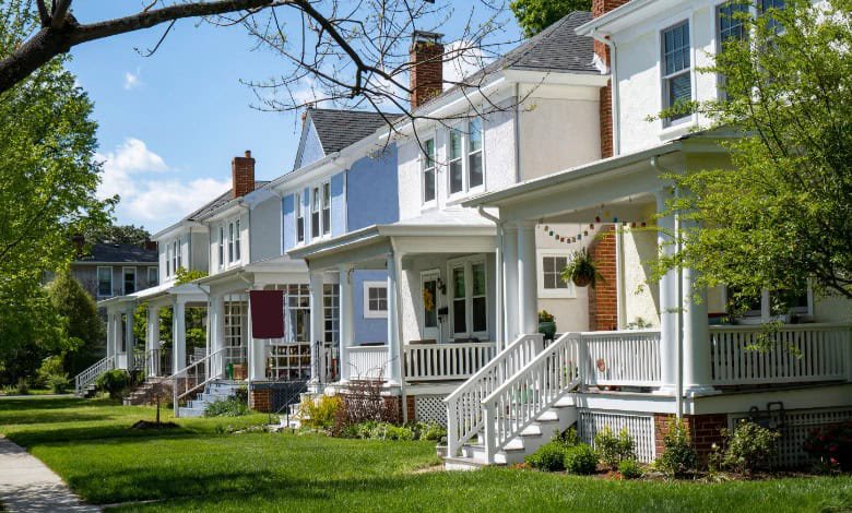 A row of older two-story houses with covered front porches, white railings, green lawns, and leafy trees along a sidewalk.